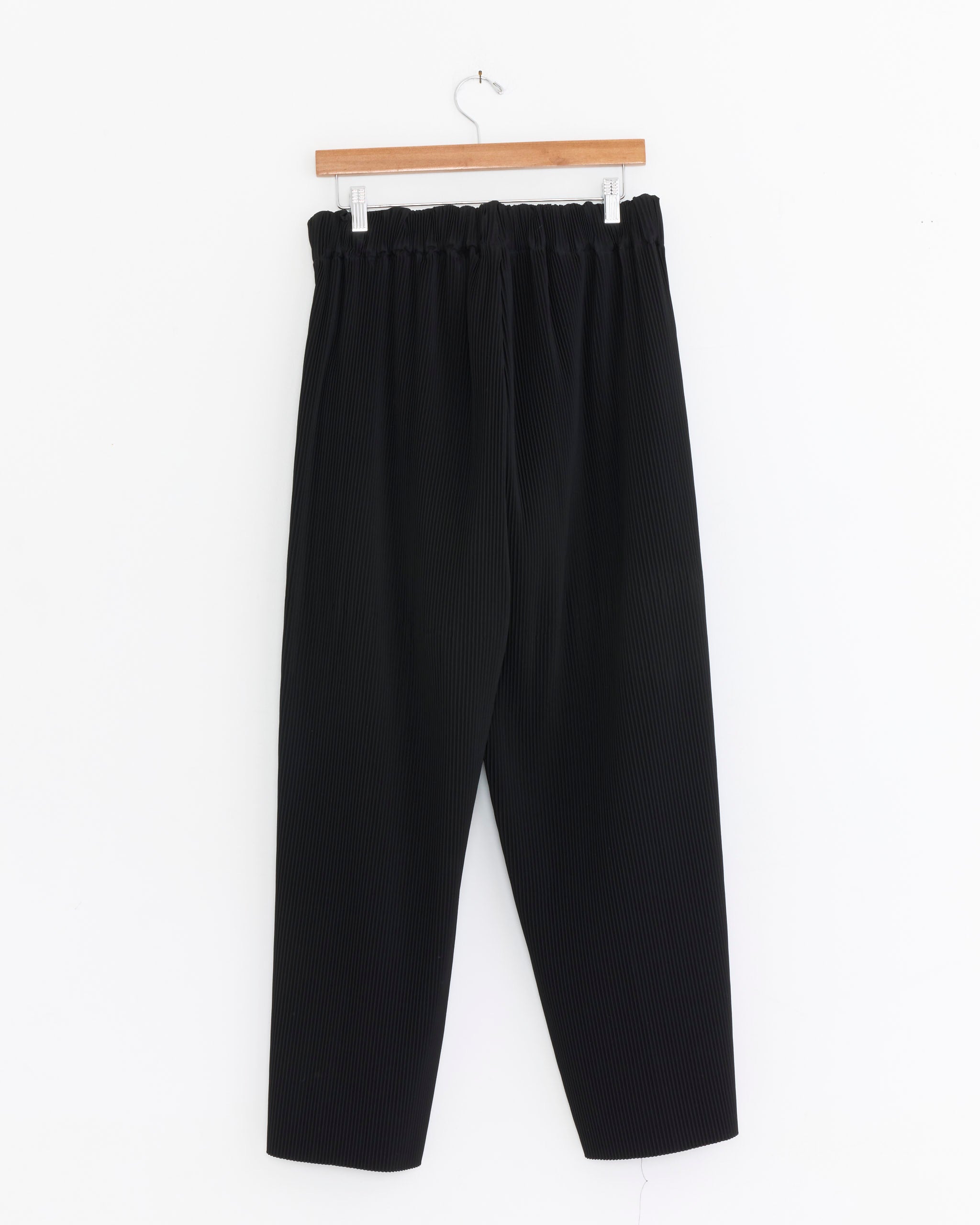 The Sillage Hakama Pockets Pleat Pant in Black, featuring an elastic waistband and wide pleated legs, hangs on a wooden hanger against a white background.