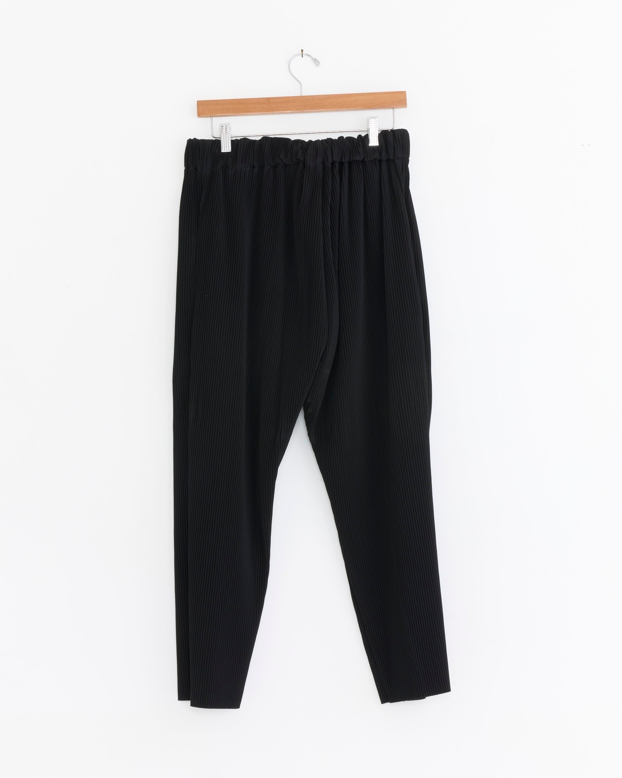 The Sillage Baggy Pockets Pleat Trouser in Black, featuring pleated details and an elastic waistband, hangs on a wooden hanger against a plain white background.