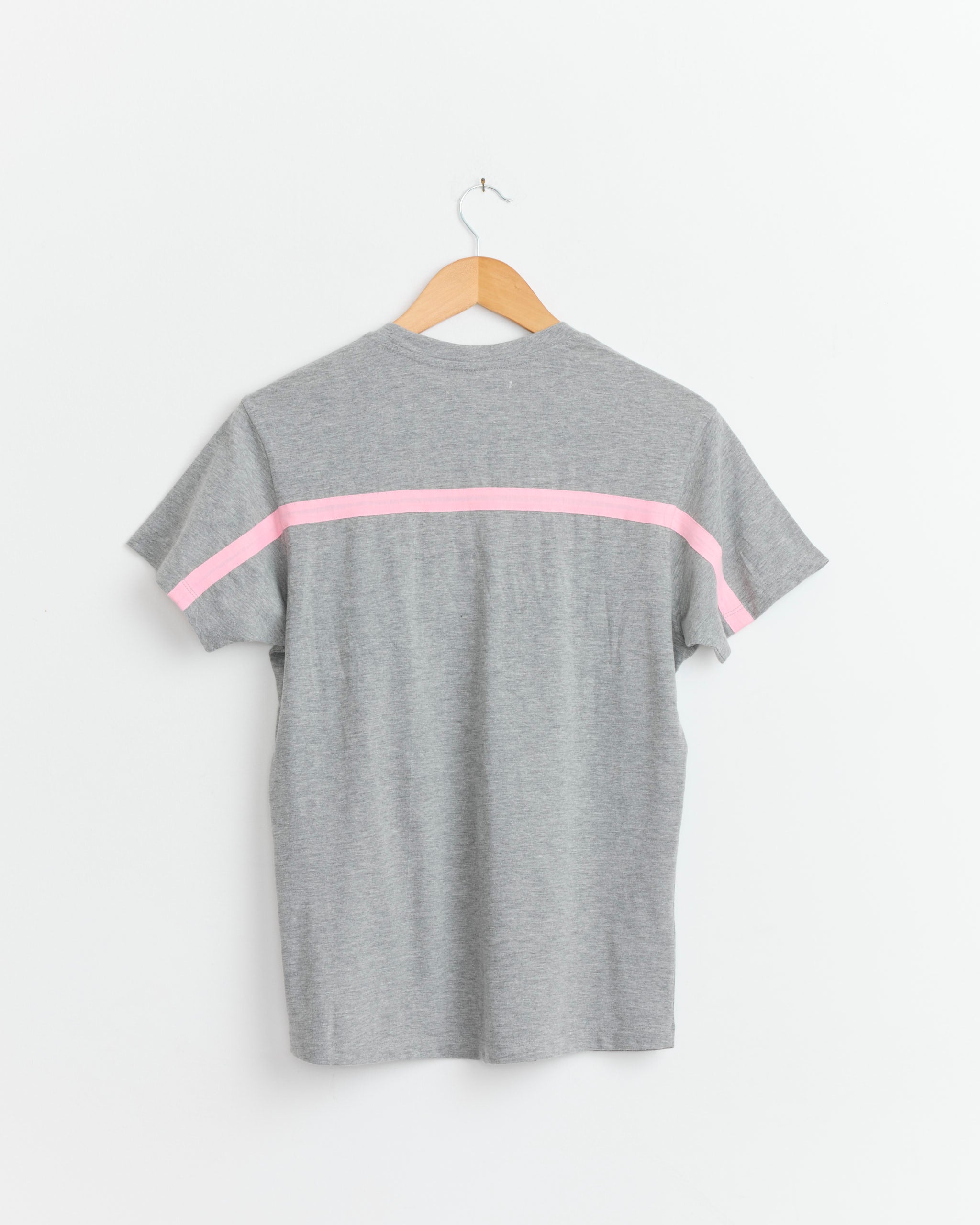 The Thomas T-Shirt in Grey by Gimaguas is a unisex tee with a relaxed fit, displayed on a wooden hanger against a white wall, and features a single pink horizontal stripe across the back of the sleeves and upper back.