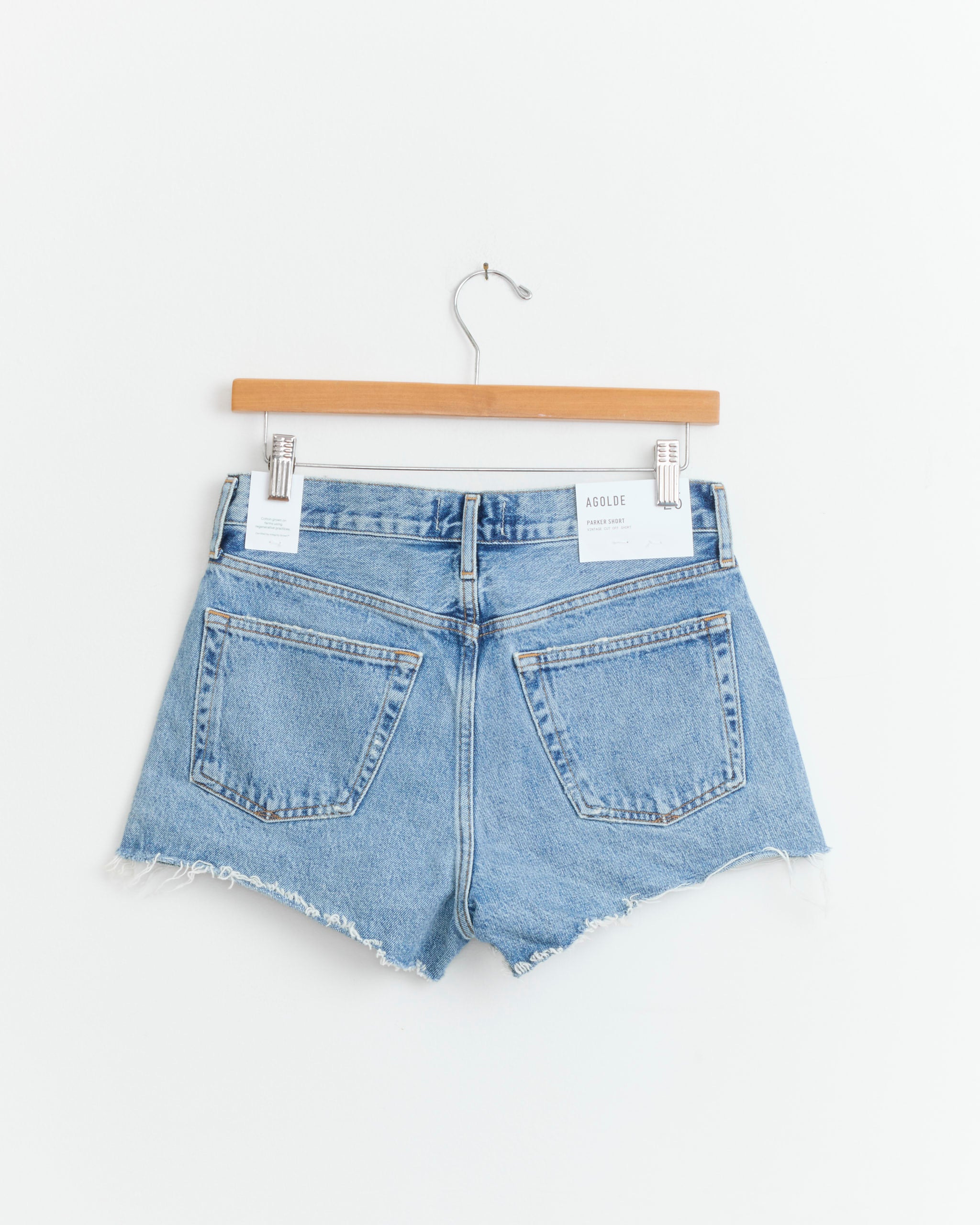 The AGOLDE Parker Short in Chorus features high-rise, light blue vintage indigo denim with frayed hems, shown on a wooden hanger against a white background.