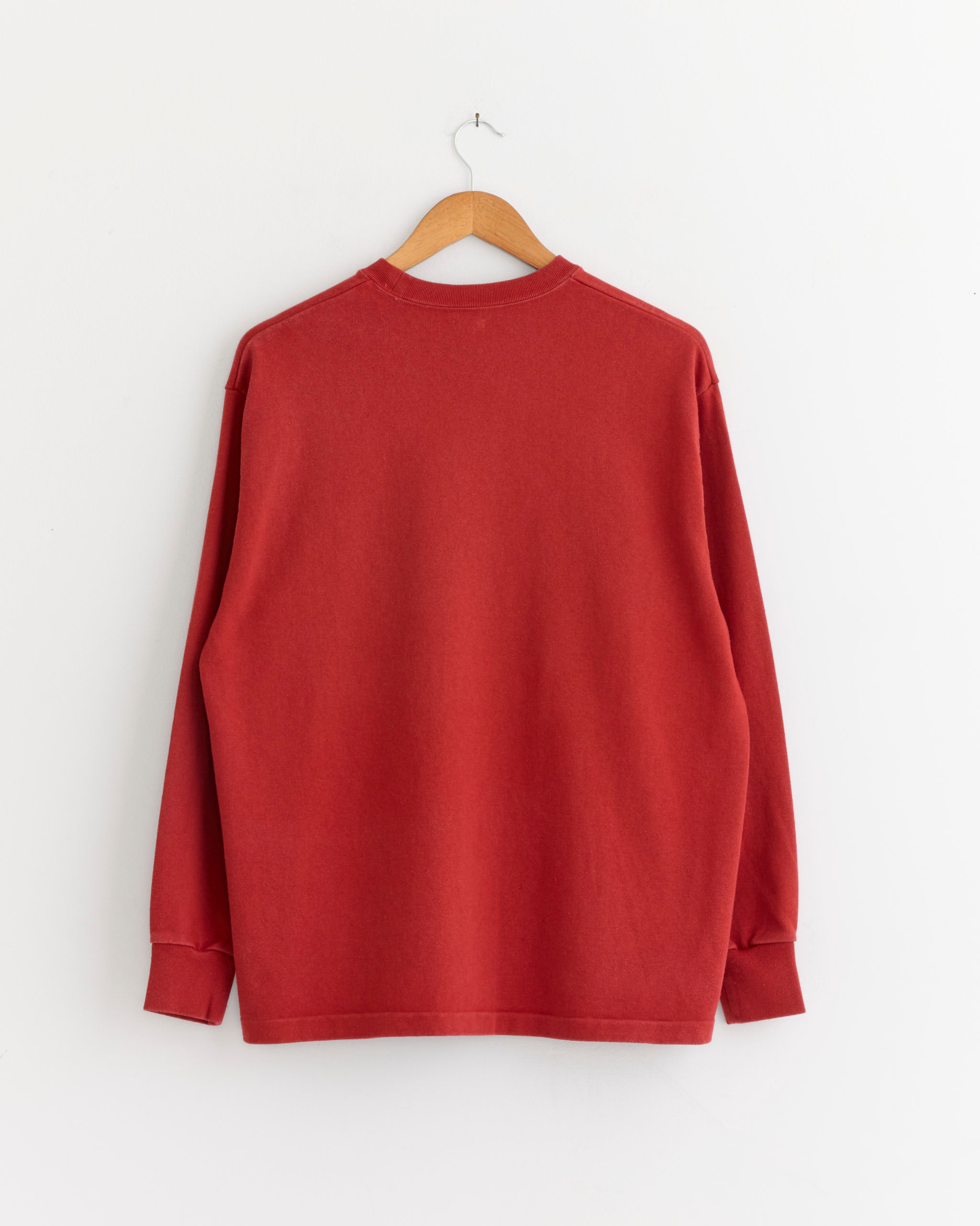 The A.PRESSE Vintage Long Sleeve T-Shirt in Red hangs on a wooden hanger against a white wall, displayed from the back.