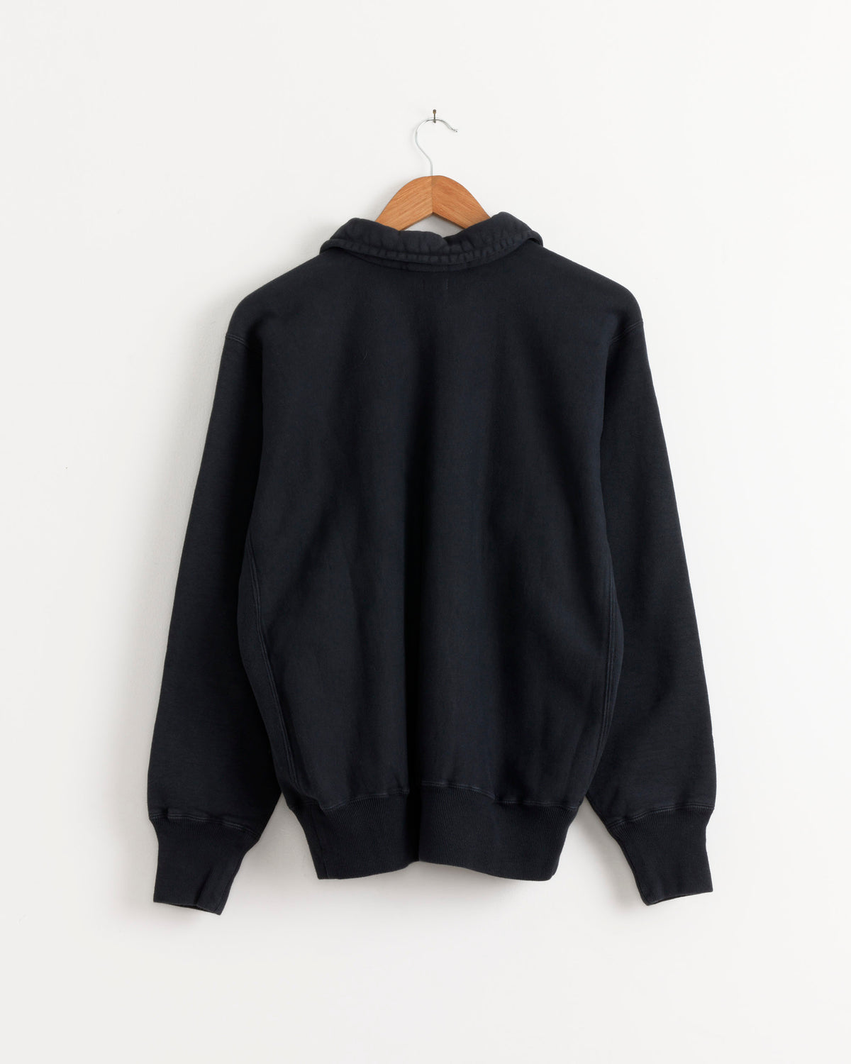 Back view of a black A.PRESSE Vintage Half Zip Sweatshirt in brushed-back cotton fleece, hanging on a wooden hanger against a white wall.