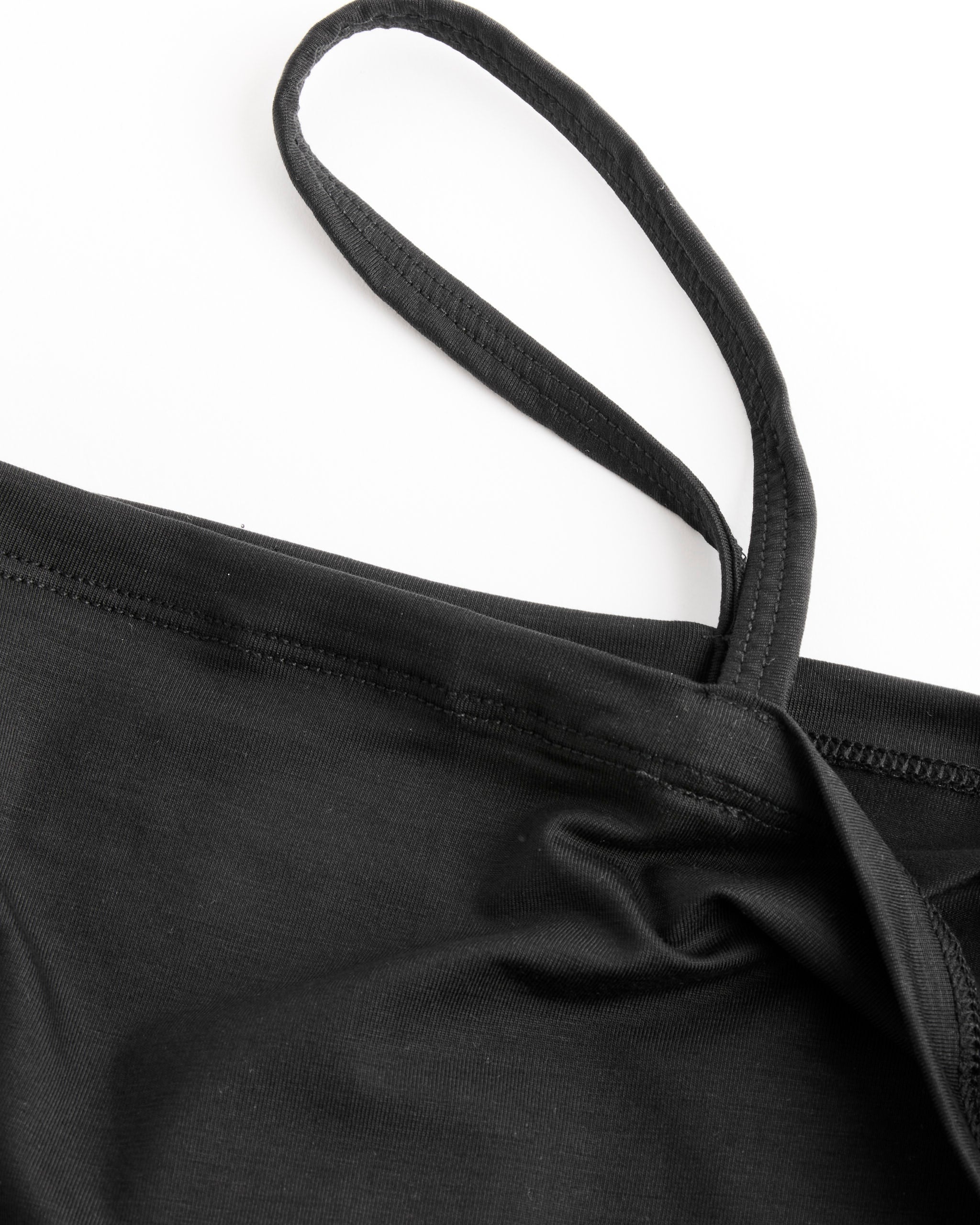 Close-up of the Baserange Nida One Strap in Black, a bamboo lyocell garment blended with elastane for stretch, featuring a visible strap and seam. The piece is laid out on a white background with slight wrinkling near the seam and strap.