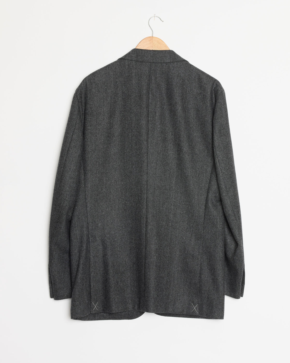 A light grey unlined blazer by Salon C. Lundman hangs on a wooden hanger against a white wall, displayed from the back with long sleeves and a straight hem. Crafted from a luxurious wool-cashmere blend, the Herringbone SB Unlined Jacket showcases timeless style and understated elegance.