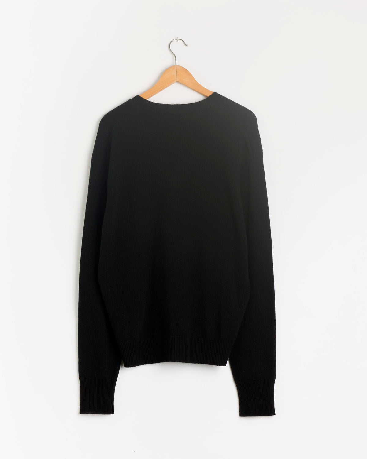The Medium Gauge V-Neck Sweater in Black by Salon C. Lundman hangs on a wooden hanger against a white wall, displayed with the back facing forward.