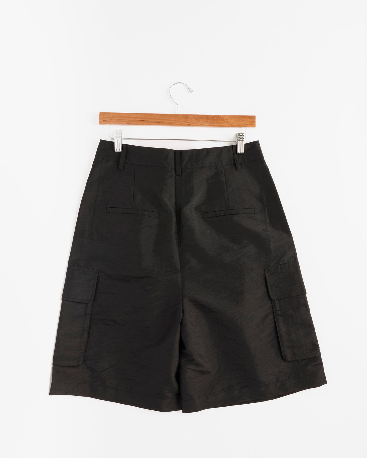 The Tibi Crispy Nylon Stella Short in Black, crafted from lightweight Italian nylon and featuring a modern knee-length cargo silhouette with front pockets, hangs on a wooden hanger against a plain white background.