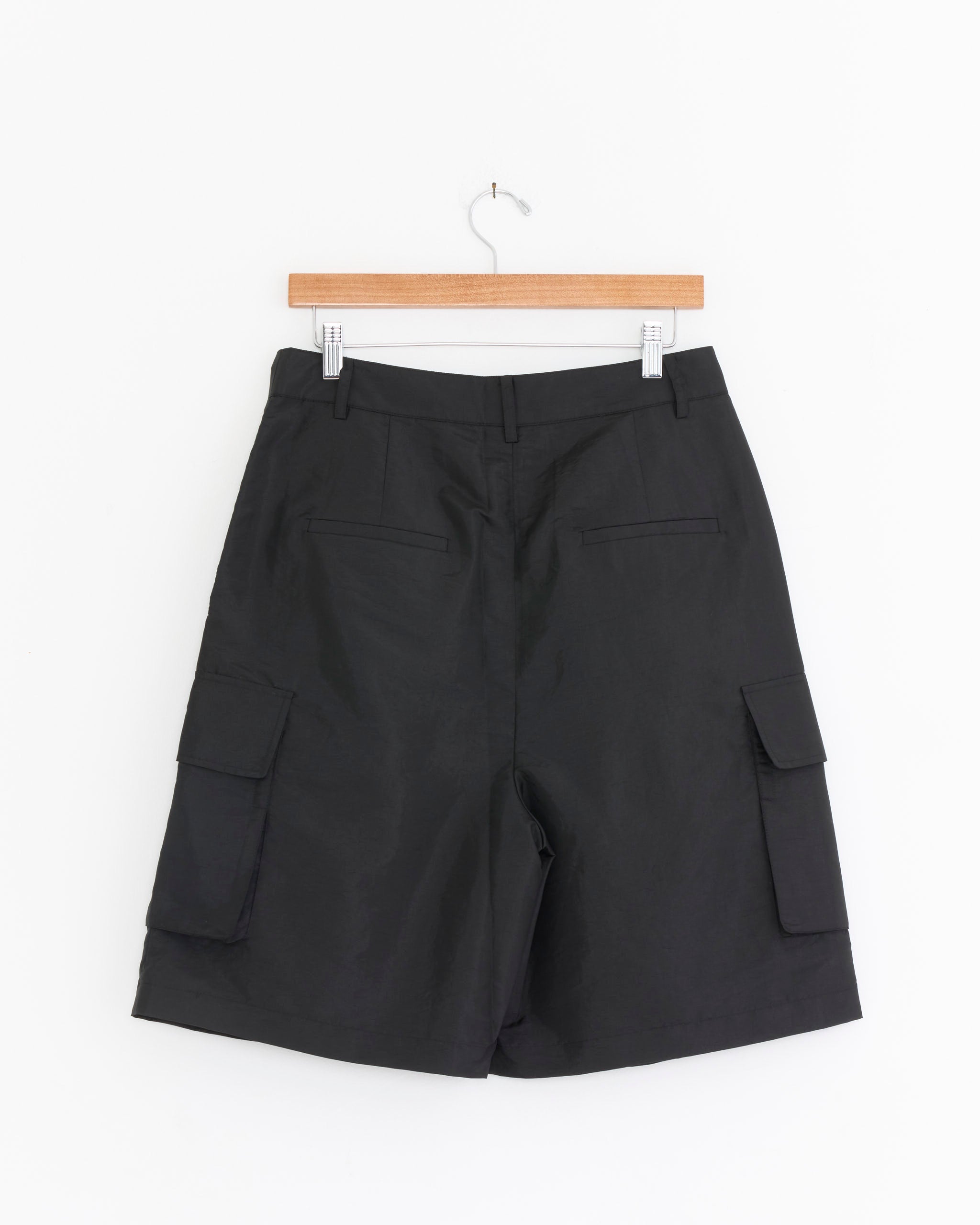 The Tibi Crispy Nylon Stella Short in Black, shown on a wooden hanger against a white background, is made from lightweight Italian nylon and features two large side cargo pockets plus two back pockets.