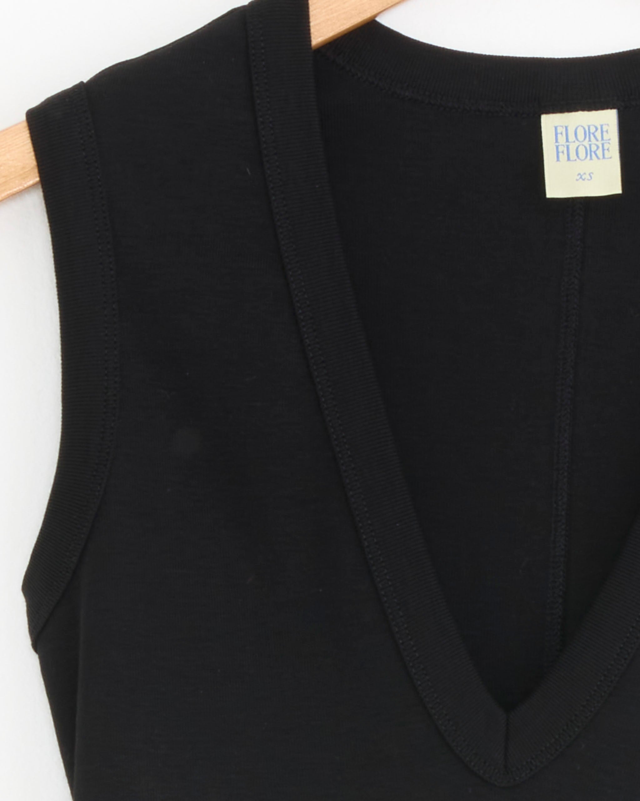 Close-up of the Dewi Tank in Black by FLORE FLORE, a sleeveless V-neck top on a wooden hanger, featuring a small light yellow label with the brand name and XS at the neckline.