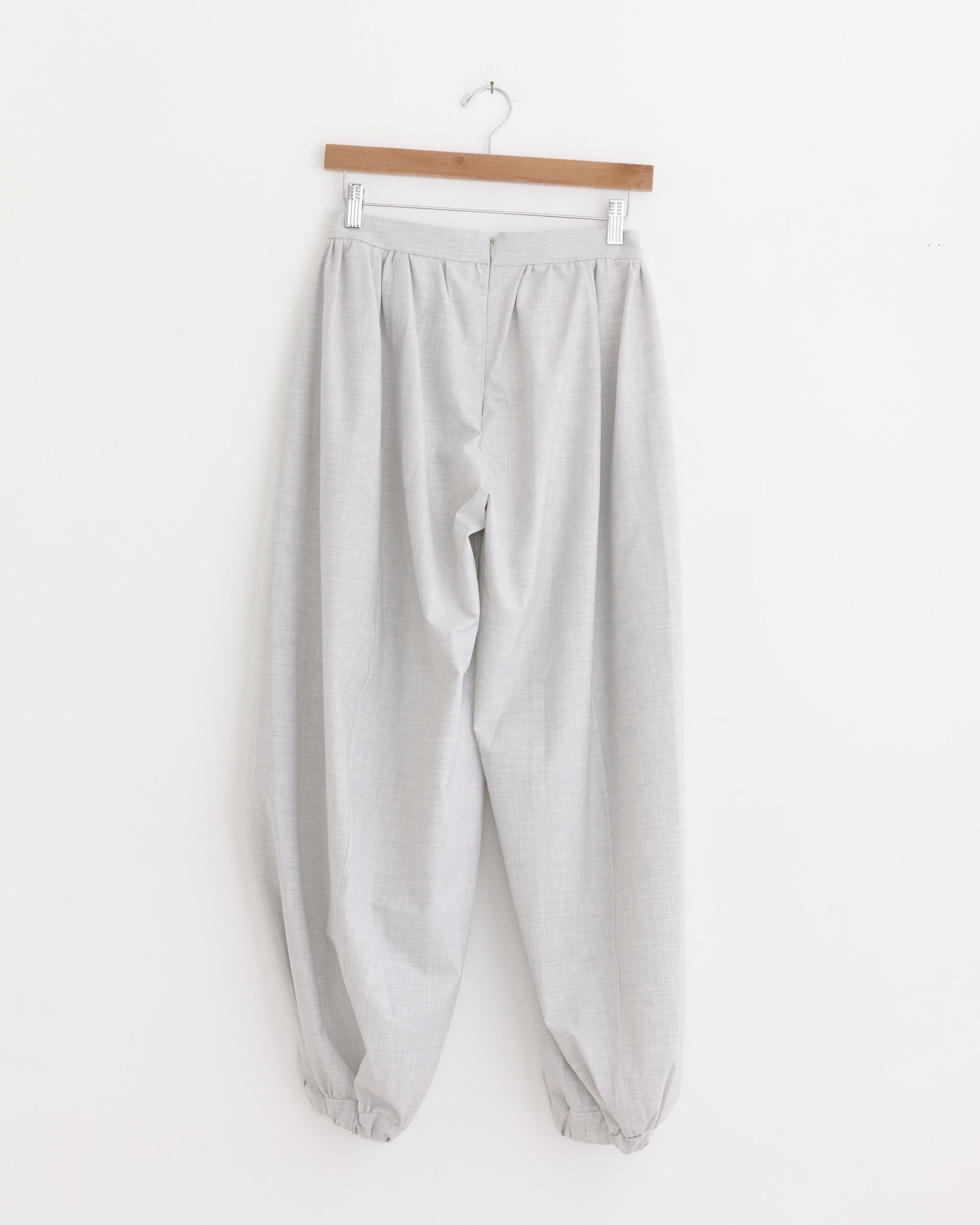 The Lucia Pant in Grey Set by Michelle Del Rio hangs on a wooden hanger against a plain white wall. This light gray, loose-fitting style showcases sculptural tailoring, an elasticated ankle, subtle texture, and a high waist for elevated everyday wear. Handcrafted in Paris.