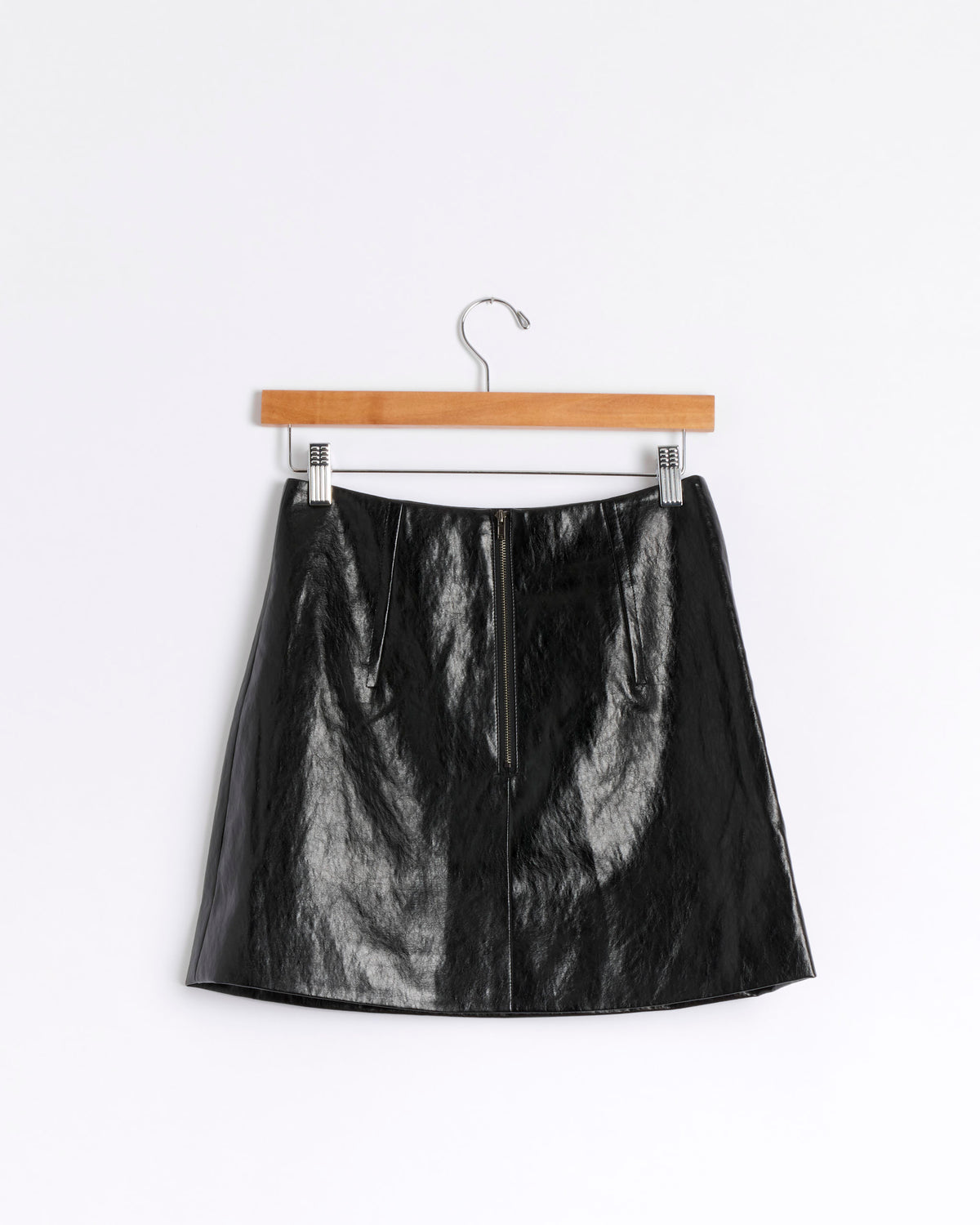 The Mijeong Park Faux Leather Mini Skirt in Black, crafted from crinkled faux leather, hangs on a wooden clothes hanger against a plain white background. The skirt showcases a visible vertical zipper down the center front.