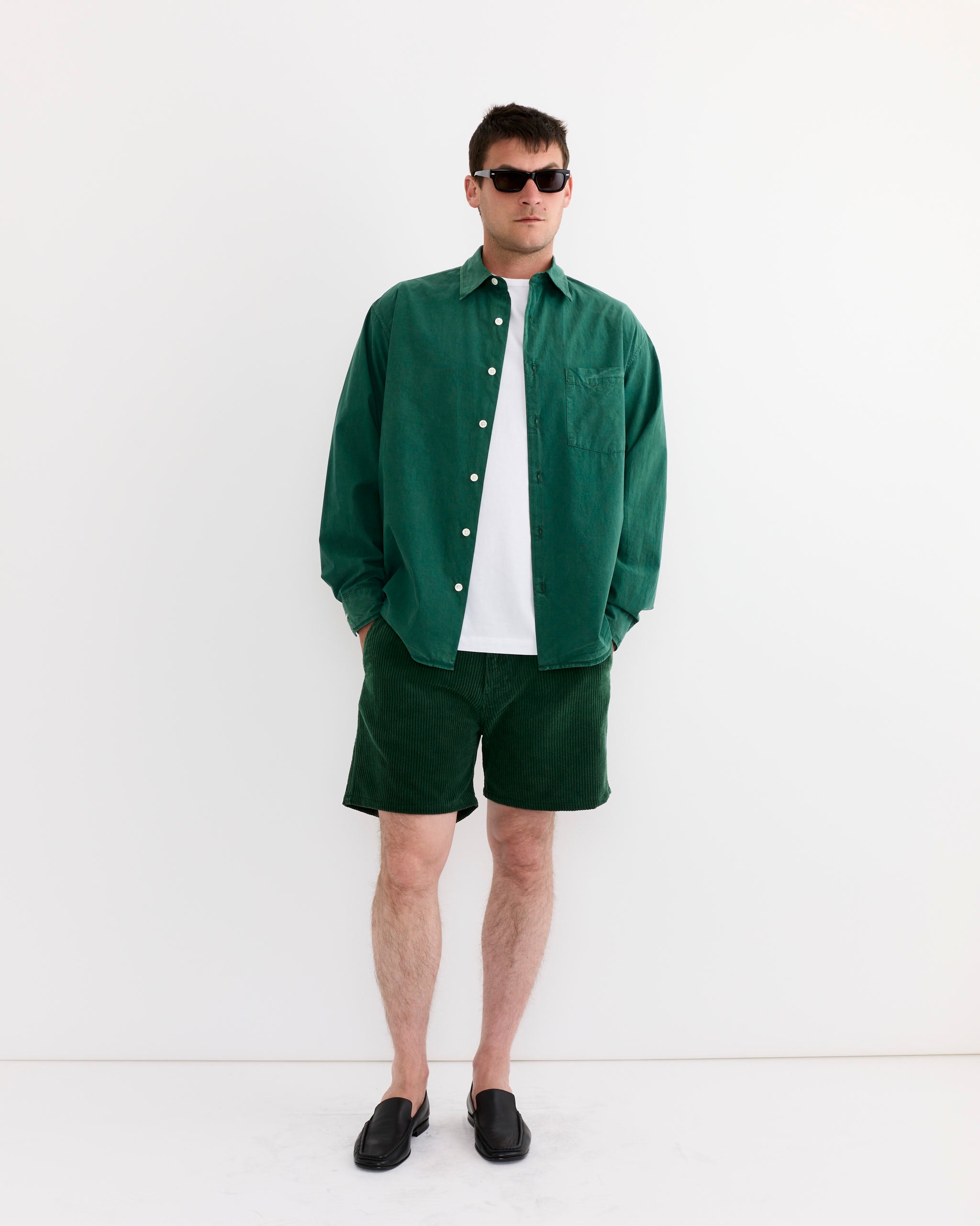 A man wears the SMOCK Routine Shirt in Cotton Silk Washed Green with matching shorts, a white tee, black sunglasses, and black slip-on shoes, standing against a plain white wall.
