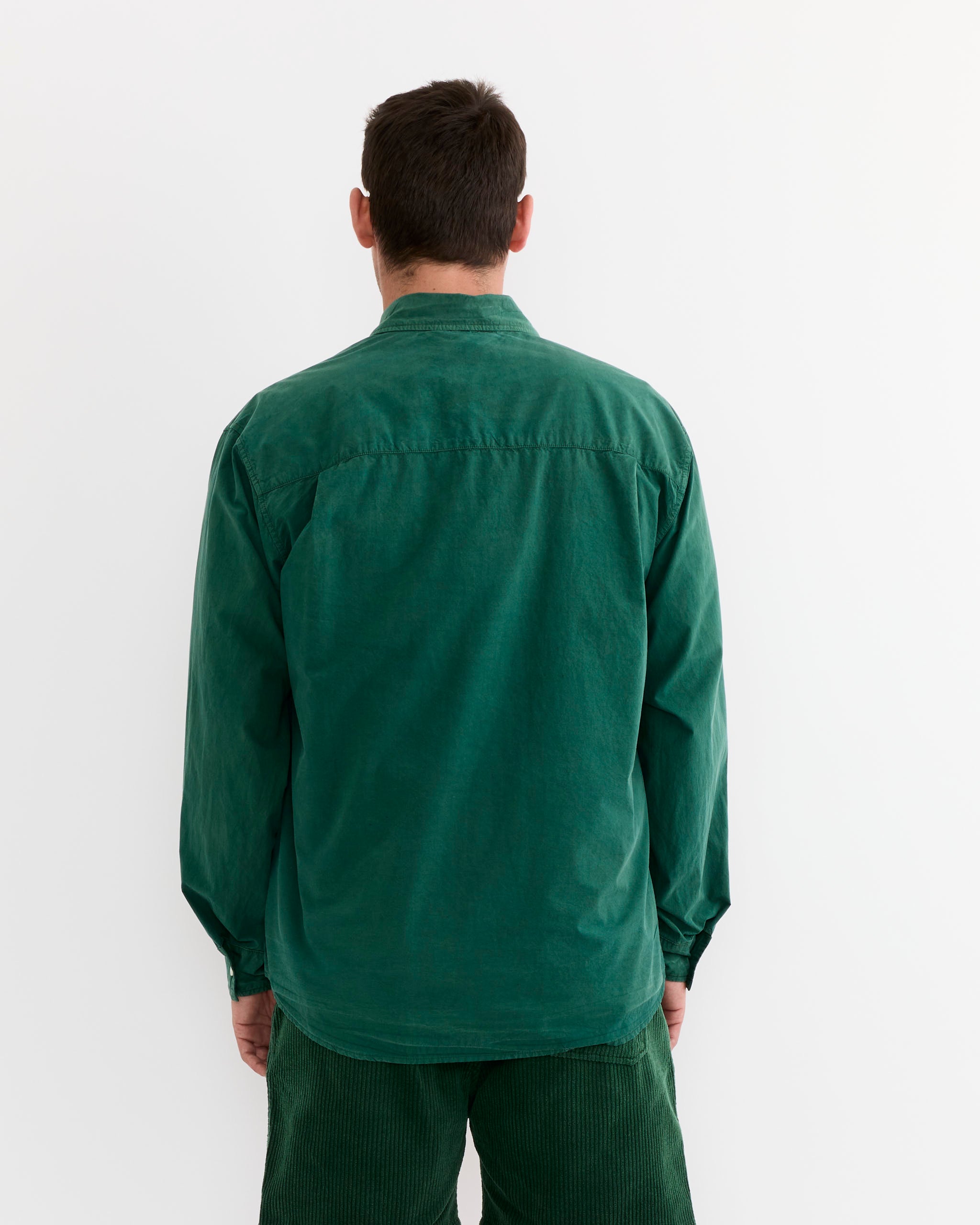 A person with short dark hair stands with their back to the camera, wearing the SMOCK Routine Shirt in Cotton Silk Washed Green, paired with green corduroy pants against a plain white background.