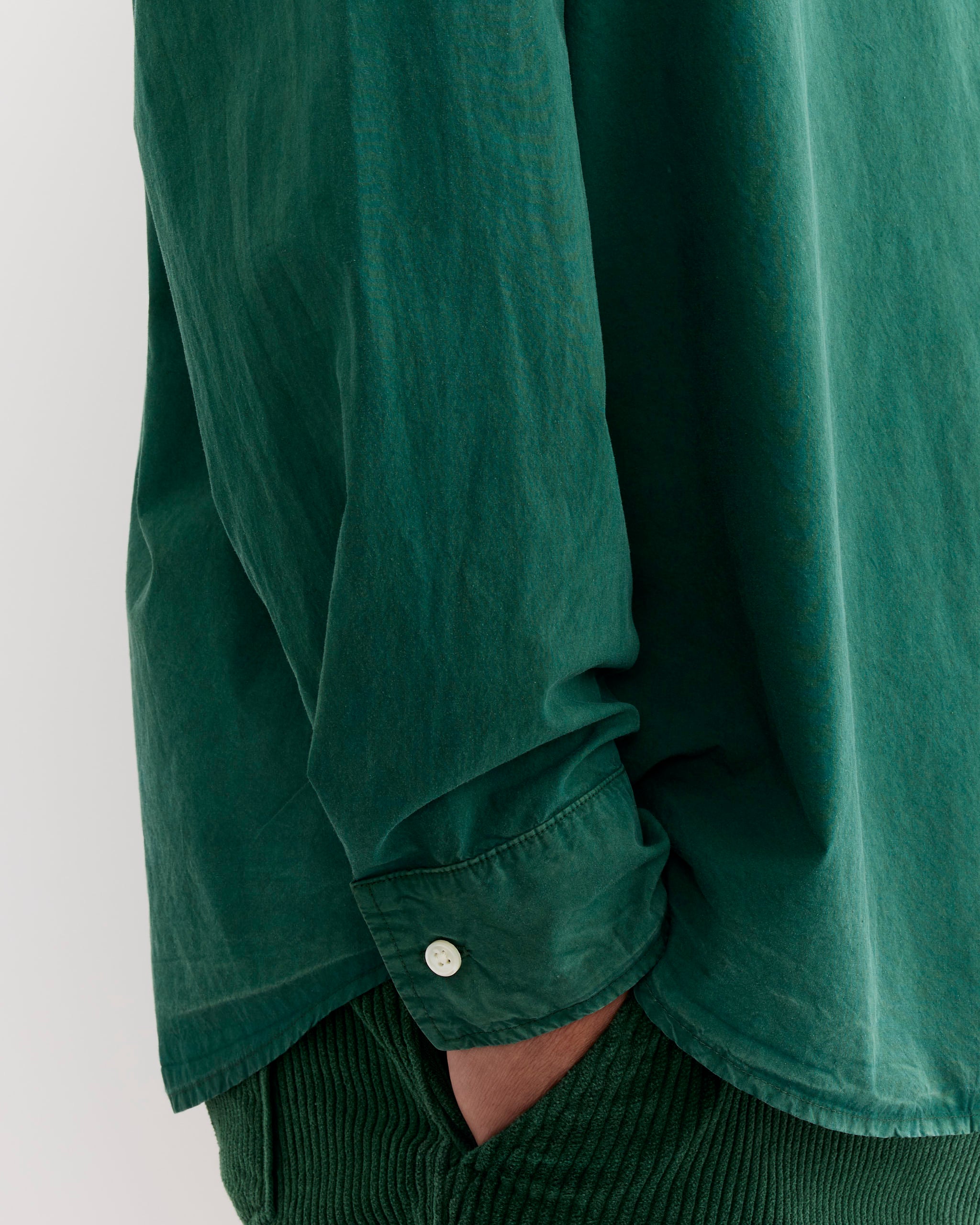 A person wears the SMOCK Routine Shirt in Cotton Silk Washed Green—loose, long-sleeved, cuffed—paired with matching green corduroy pants. The cropped image shows only their torso and arm with a hand in the pant pocket.