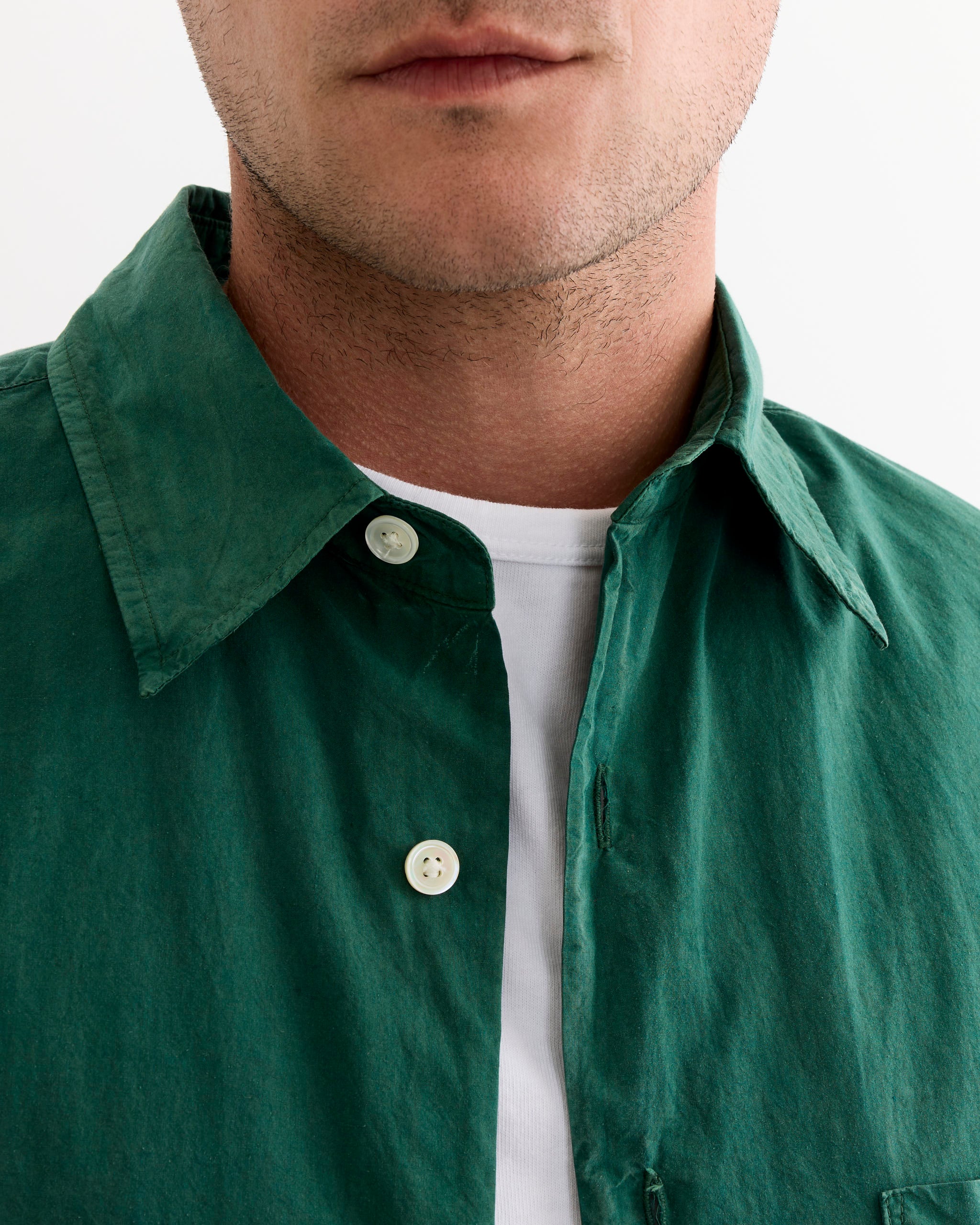 A close-up shows the lower face, neck, and upper torso of a person wearing the SMOCK Routine Shirt in Cotton Silk Washed Green, made in Los Angeles. The shirt is partially unbuttoned and layered over a white t-shirt.