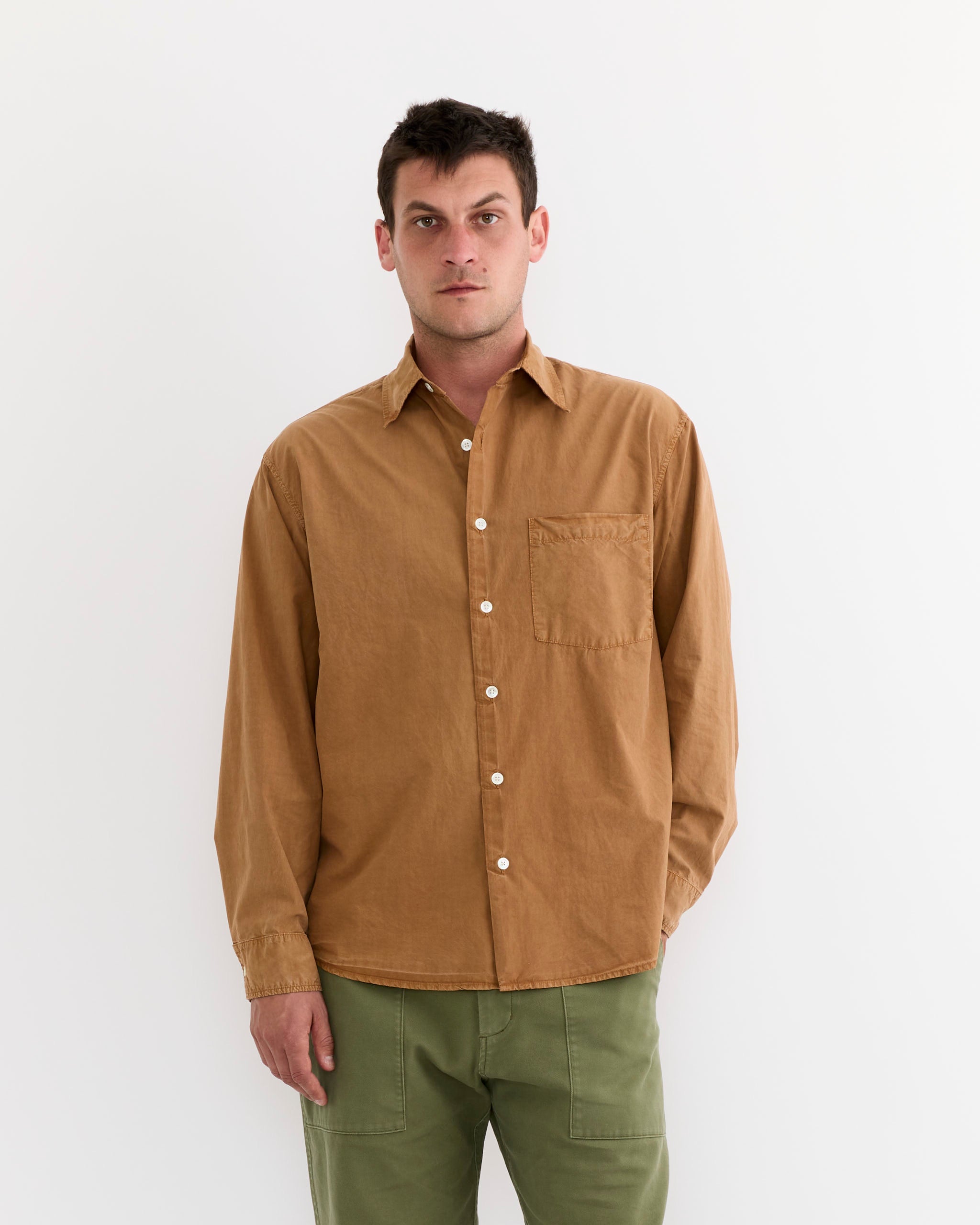 A man with short dark hair and a neutral expression stands against a white background wearing SMOCK’s Routine Shirt in Cotton Silk Washed Chestnut, featuring a chest pocket, paired with olive green pants. One hand is in his pocket.