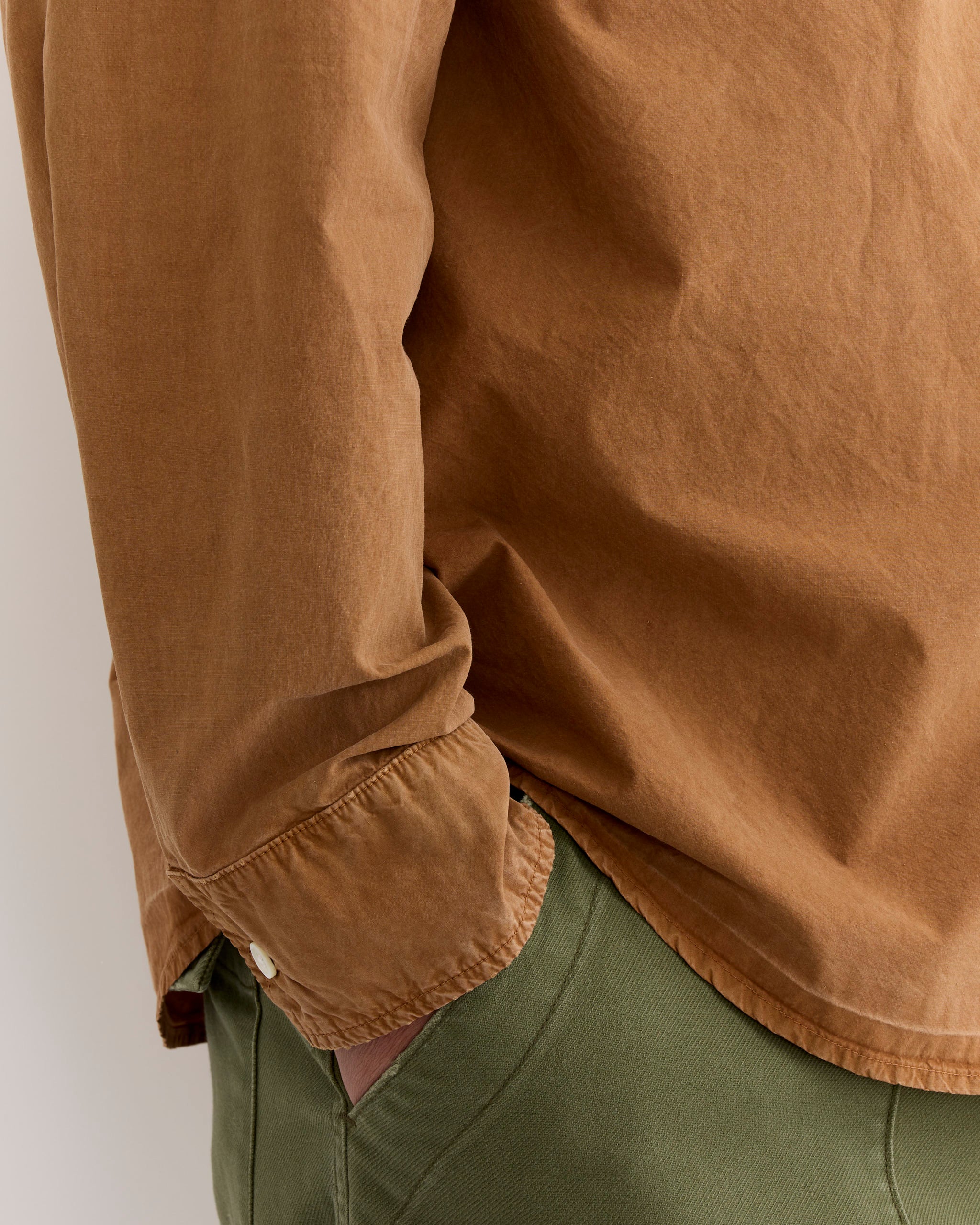 A person is seen from the side wearing SMOCK's Routine Shirt in Cotton Silk Washed Chestnut with partly rolled-up sleeves and olive green pants, one hand in their pocket.