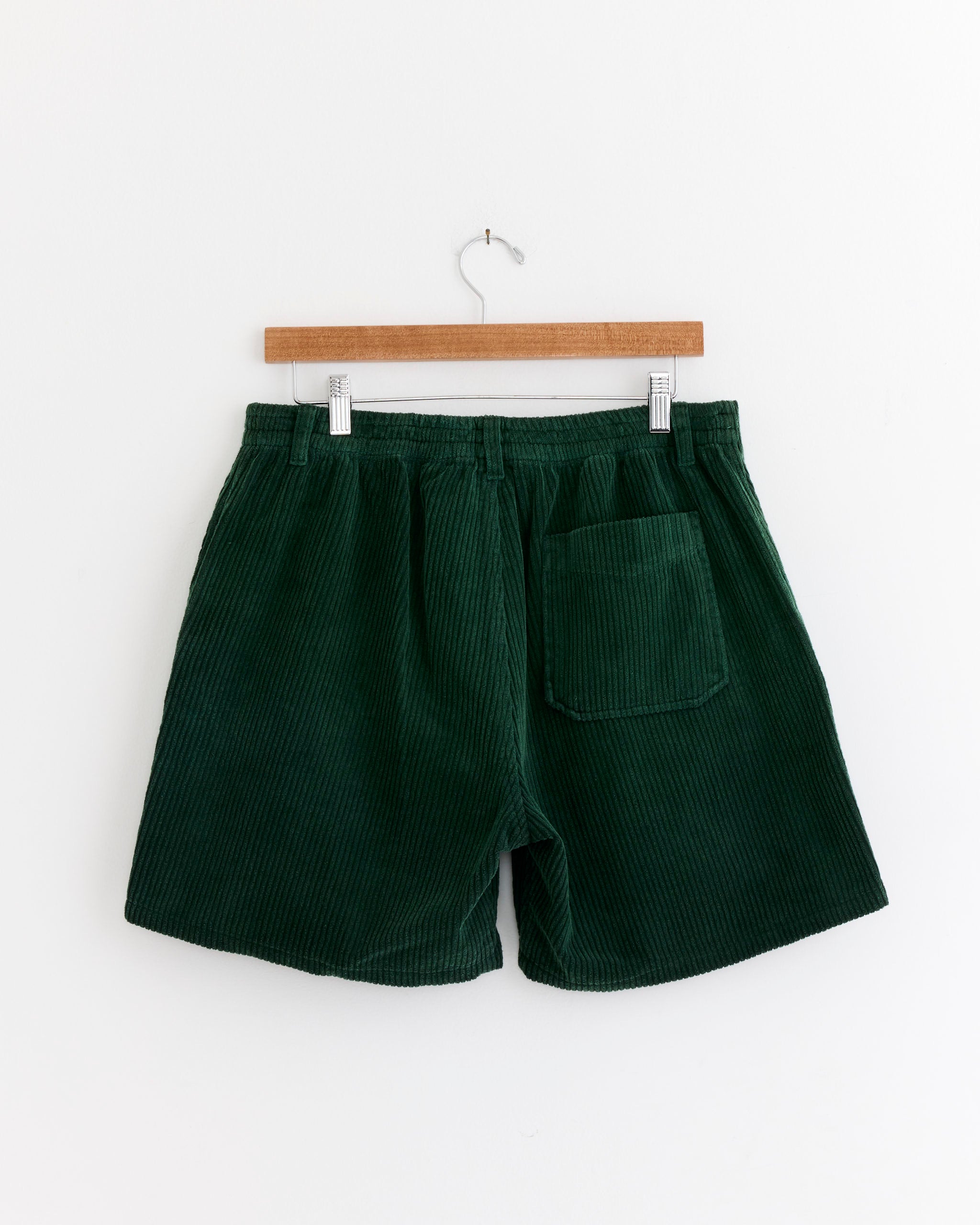 The SMOCK Laguna Short in Corduroy Washed Green features an elastic waistband, back pocket, and garment-dyed finish, shown hanging on a wooden hanger against a white wall.