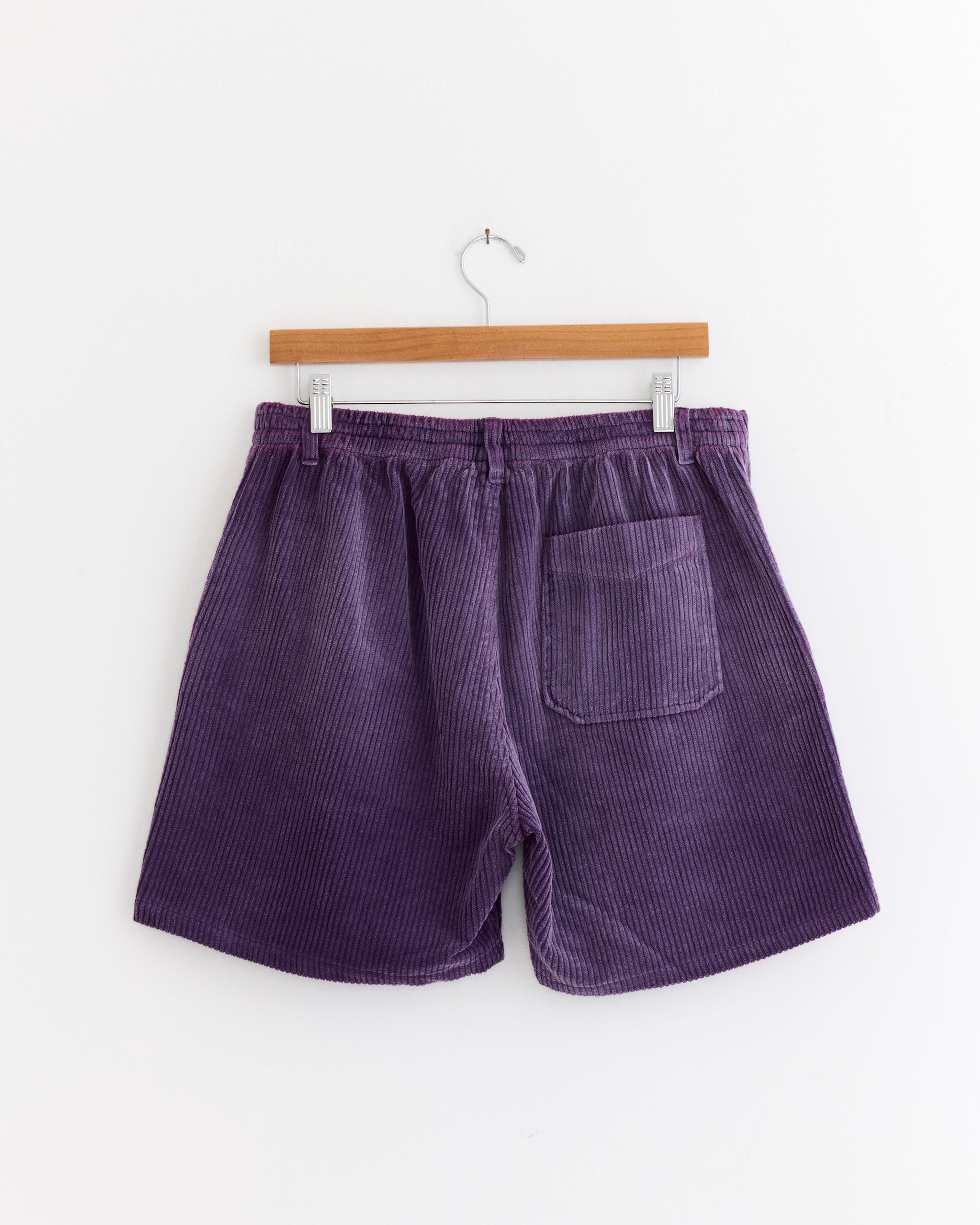 The SMOCK Laguna Short in Corduroy Washed Purple features garment-dyed cotton, an elastic waistband, and a single back pocket, shown on a wooden hanger against a white background.
