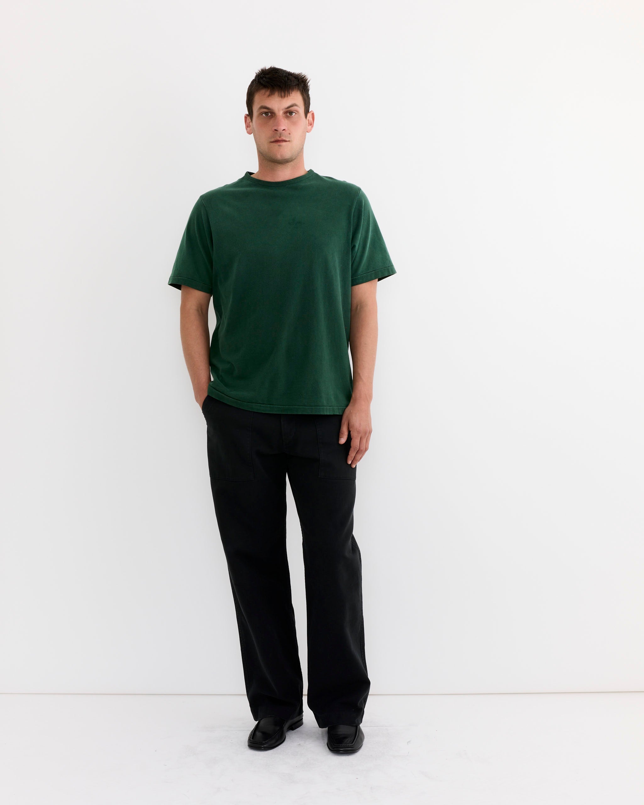 A man with short dark hair and a neutral expression stands against a plain white background, wearing the SMOCK Under Tee in Washed Green, black pants, and black shoes. His left hand is in his pocket.