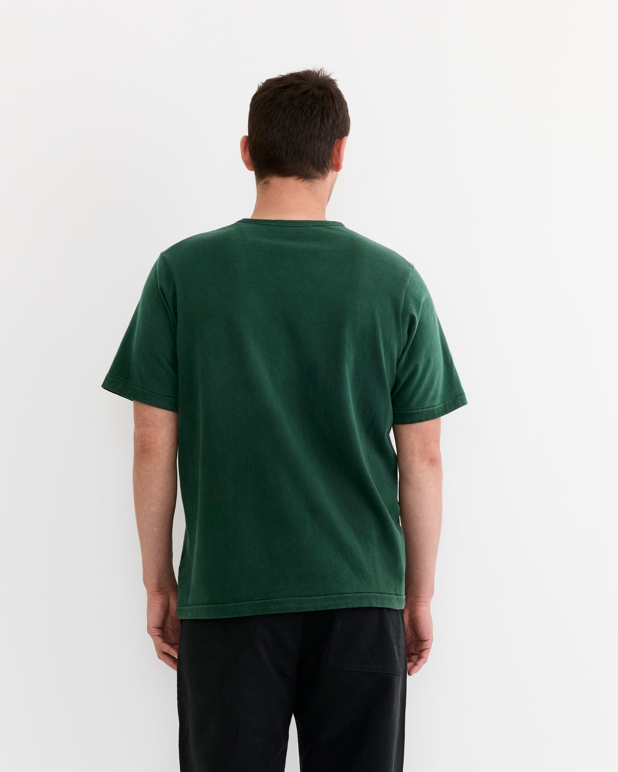 A person stands facing away from the camera against a plain white background, wearing the SMOCK Under Tee in Washed Green and black pants.