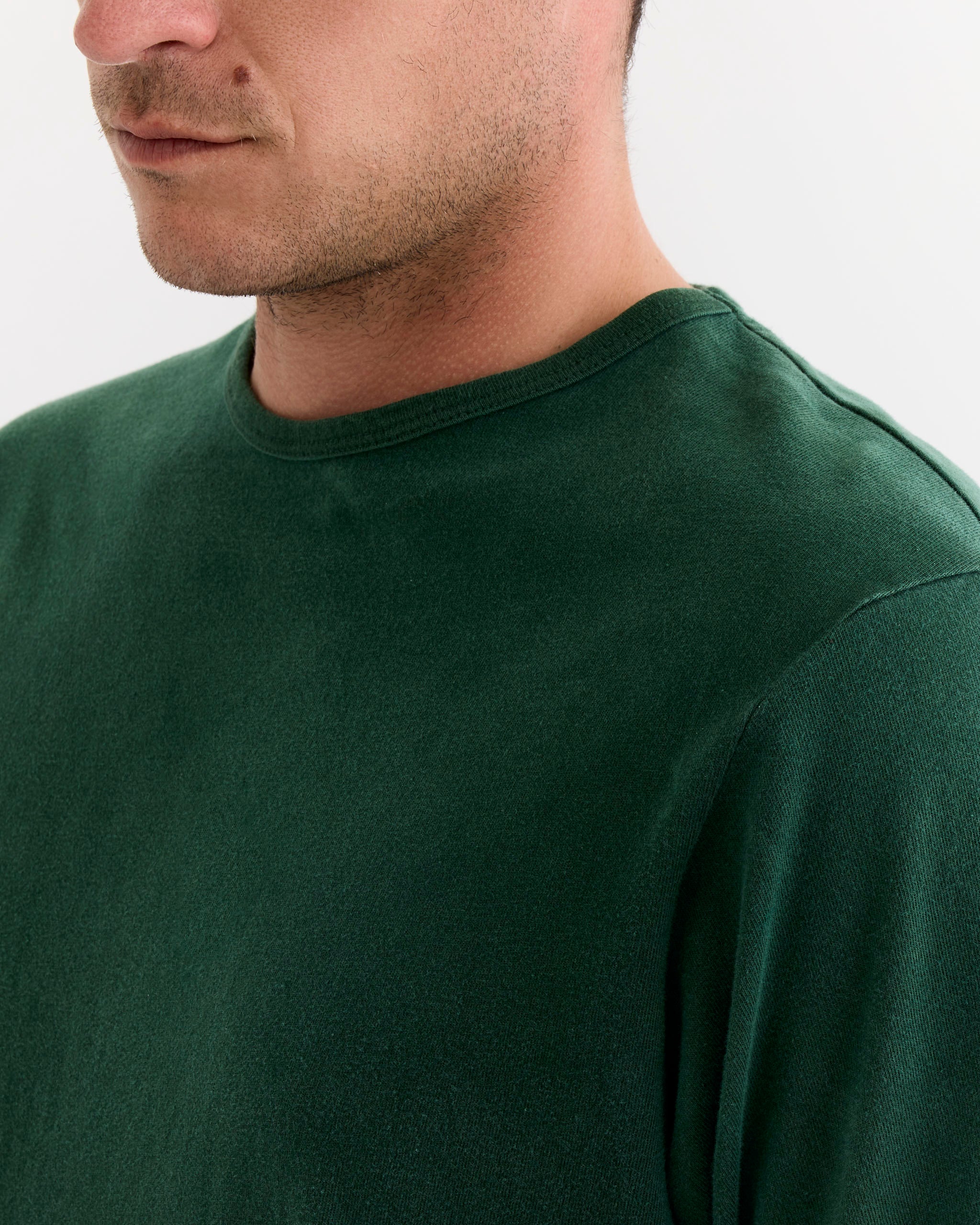 A close-up of a person wearing the SMOCK Under Tee in Washed Green, a garment-dyed cotton crew-neck—part of their face, neck, and shoulder visible against a plain light background.