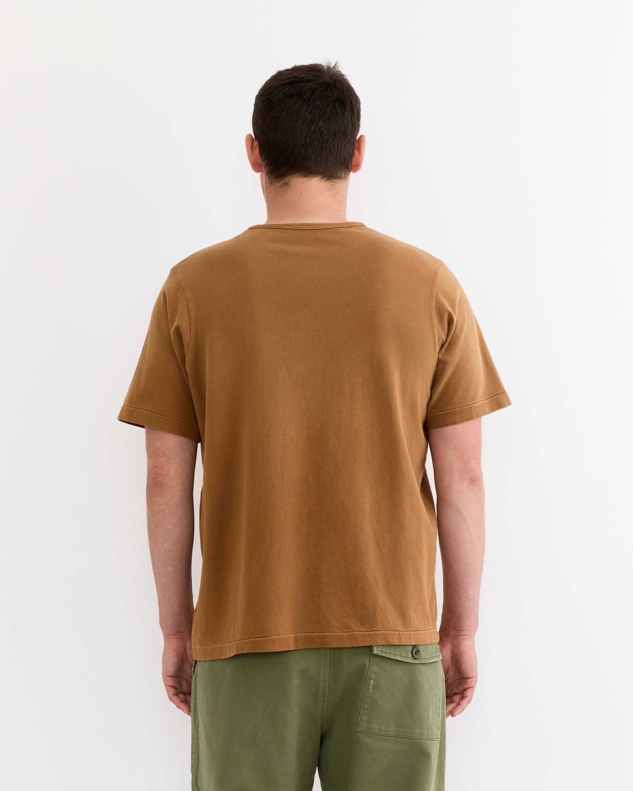 A person with short dark hair, facing away from the camera against a white background, wears the SMOCK Under Tee in Washed Chestnut, crafted from garment-dyed cotton in Los Angeles, paired with green pants.