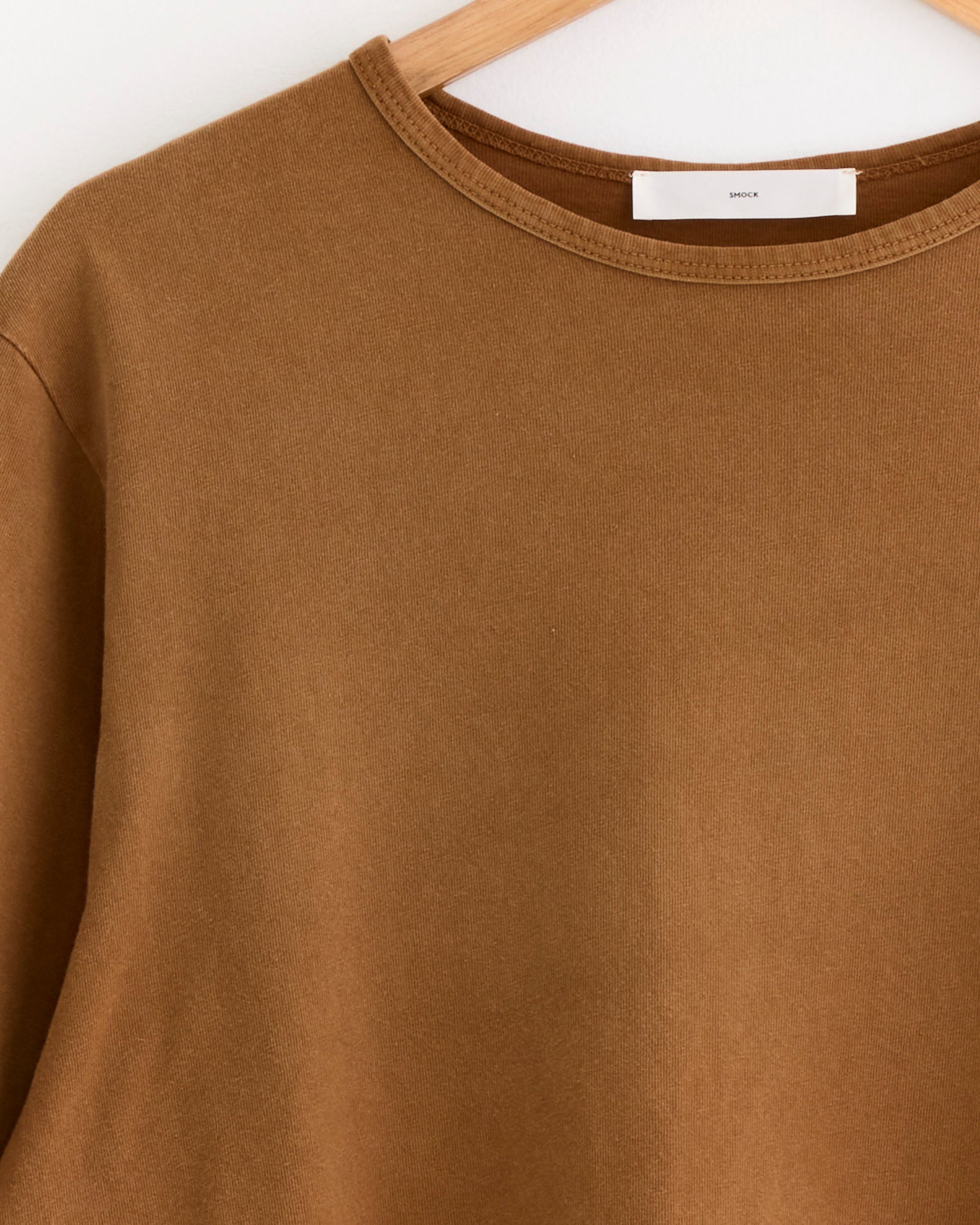 A close-up of the SMOCK Under Tee in Washed Chestnut, made from garment-dyed cotton, hangs on a wooden hanger against a white background.