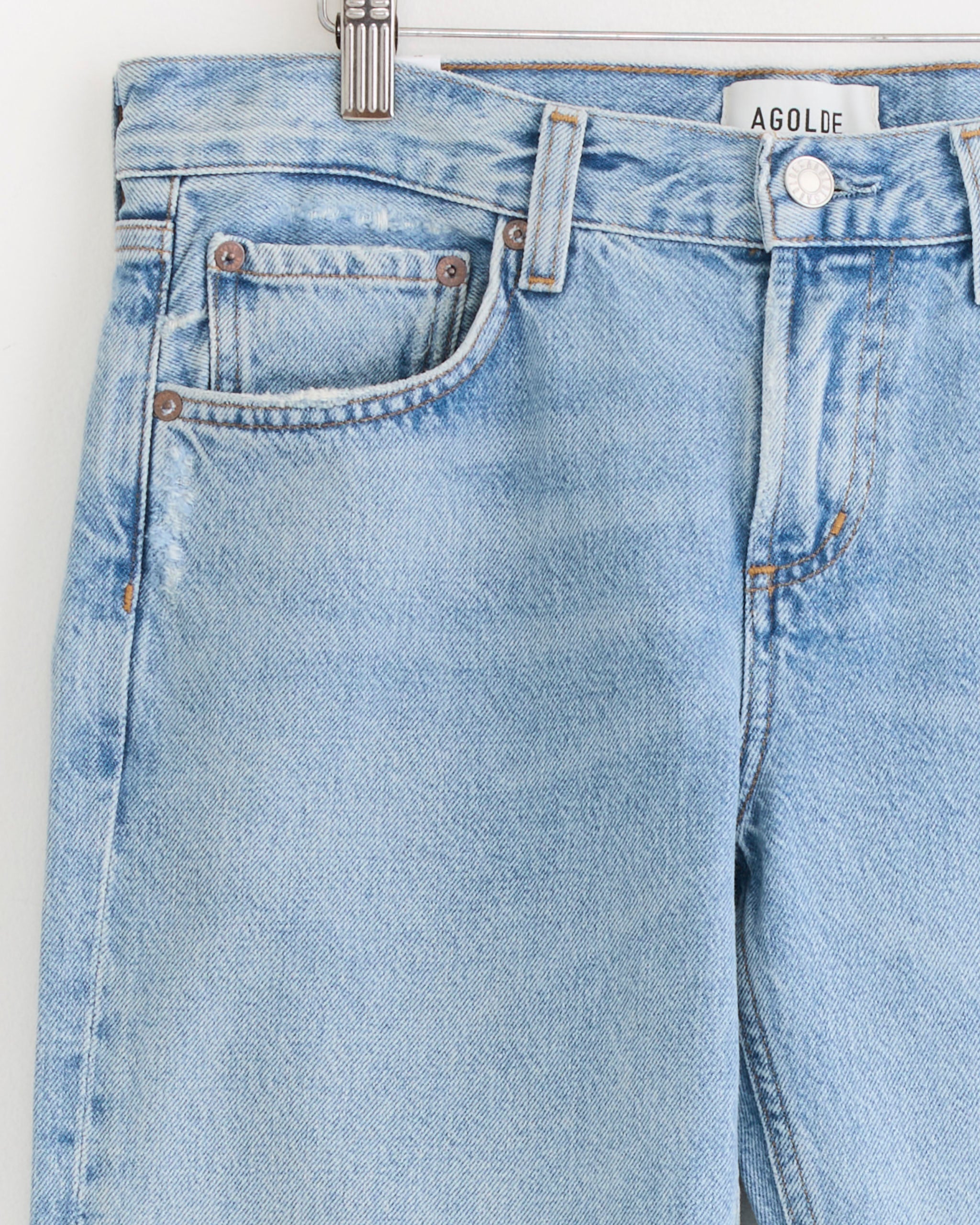 Close-up of the AGOLDE Stovepipe 29" Jean in Addition, featuring light blue denim, metal button, front pocket, and belt loops on a hanger with visible brand label. For sizing or fit help, please use our online chat.