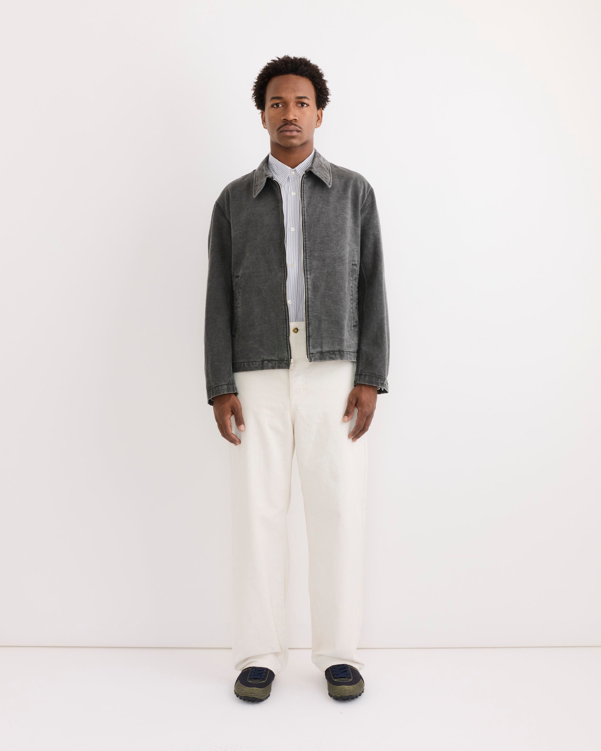A man stands against a white wall wearing the SMOCK Triple Stitch Hydra Jacket in Washed Pigment Dyed Cotton Grey, over a light blue shirt with white pants and dark shoes, facing forward to display fit for easy online sizing assistance.