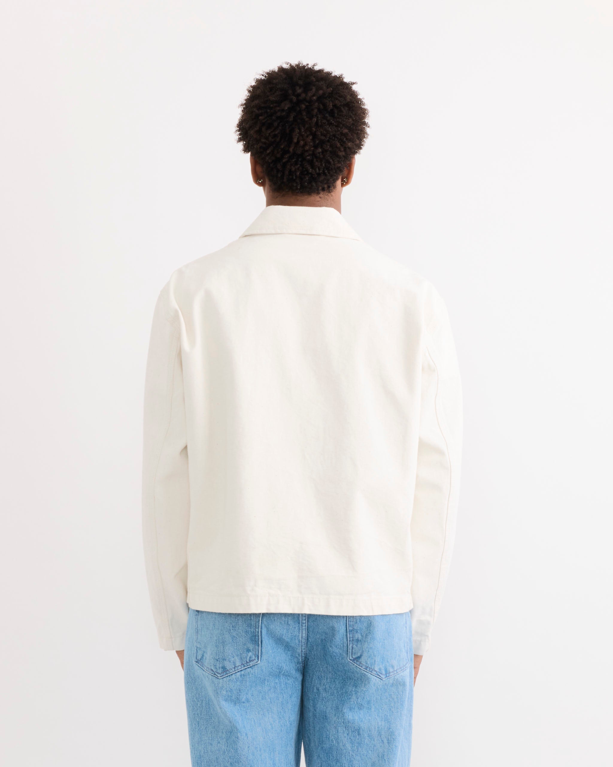 A person with short curly hair wears the SMOCK Triple Stitch Hydra Jacket in Washed Pigment Dyed Cotton Natural and light blue jeans, standing with their back to the camera against a plain white background.