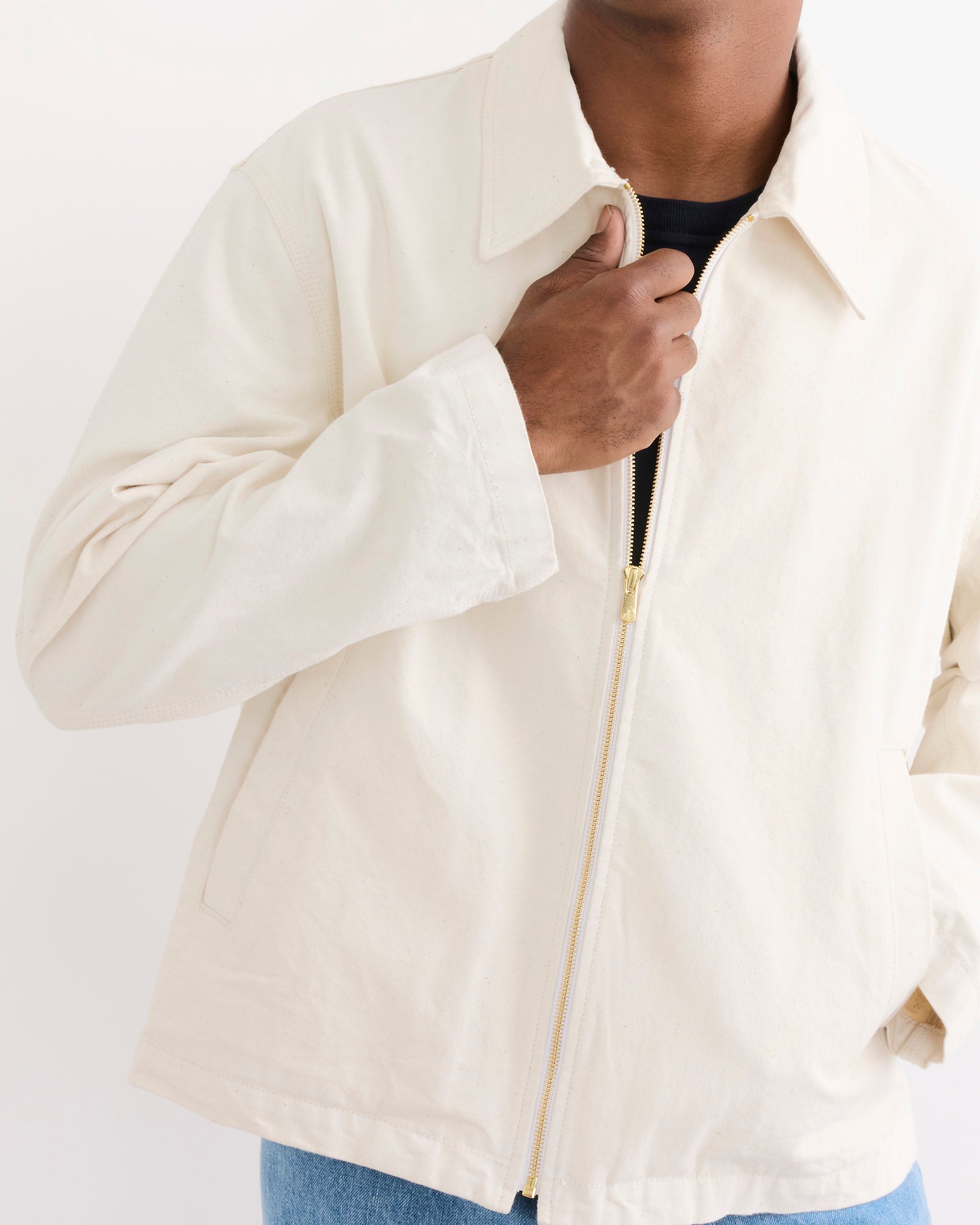 A person models the SMOCK Triple Stitch Hydra Jacket in Washed Pigment Dyed Cotton Natural over a black shirt and blue jeans, holding the collar with one hand; their head is partly out of frame to highlight fit for sizing.