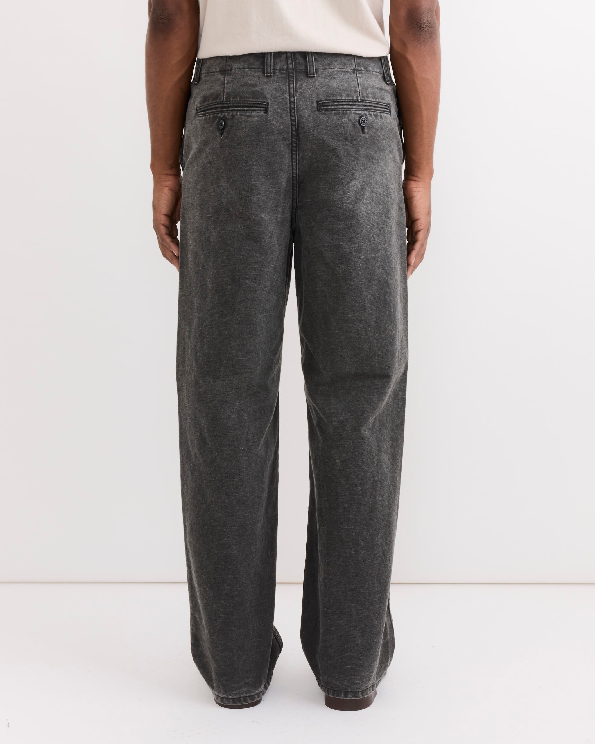 A person wearing SMOCK's Officer Trouser in Washed Pigment Dyed Cotton Grey with a relaxed fit and a light shirt stands facing away from the camera against a plain white background.