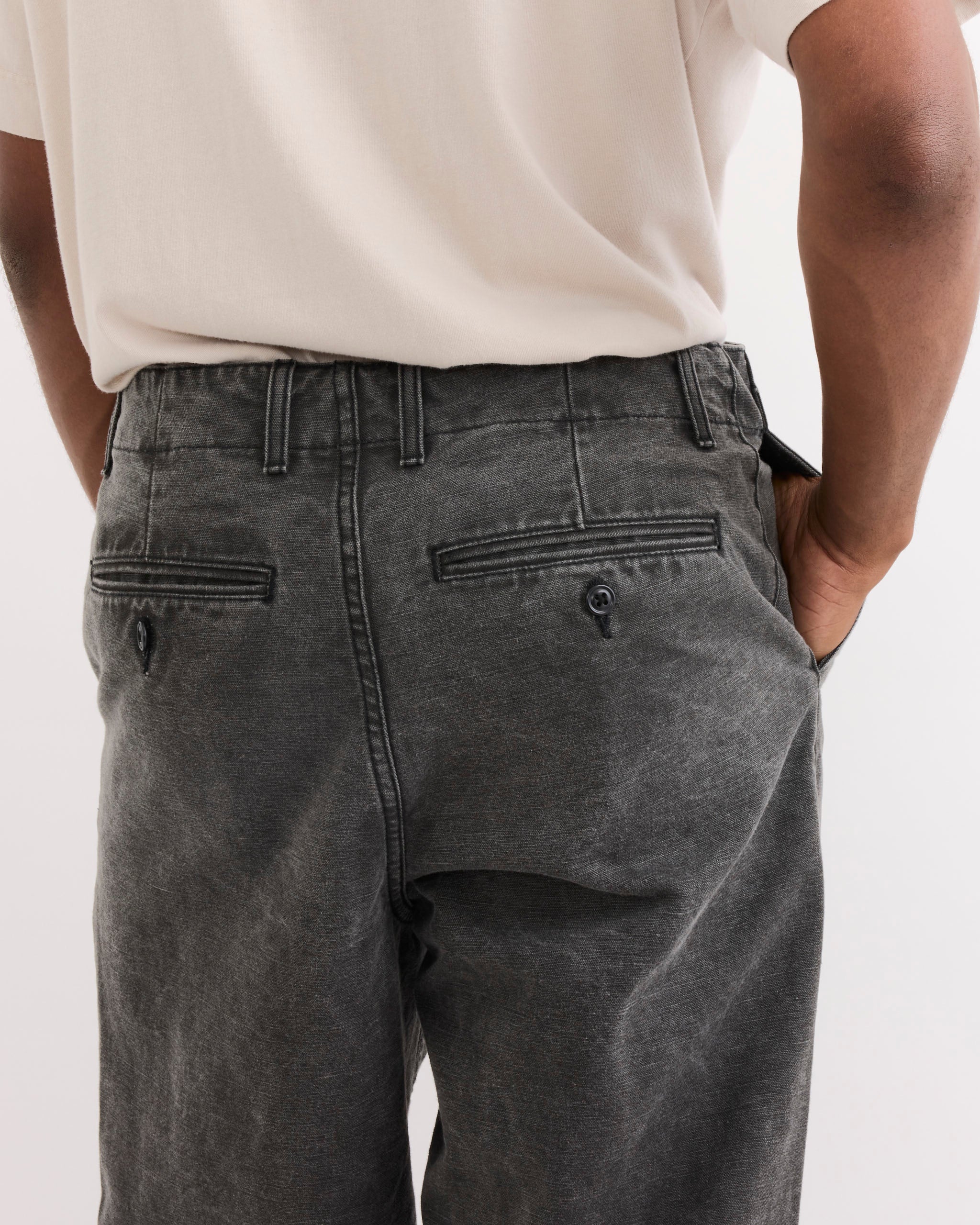 A person wearing SMOCK's Officer Trouser in Washed Pigment Dyed Cotton Grey, shown from the back with hands in pockets. The pants have belt loops, two buttoned back pockets, and a relaxed fit with an inseam just above the shoes.