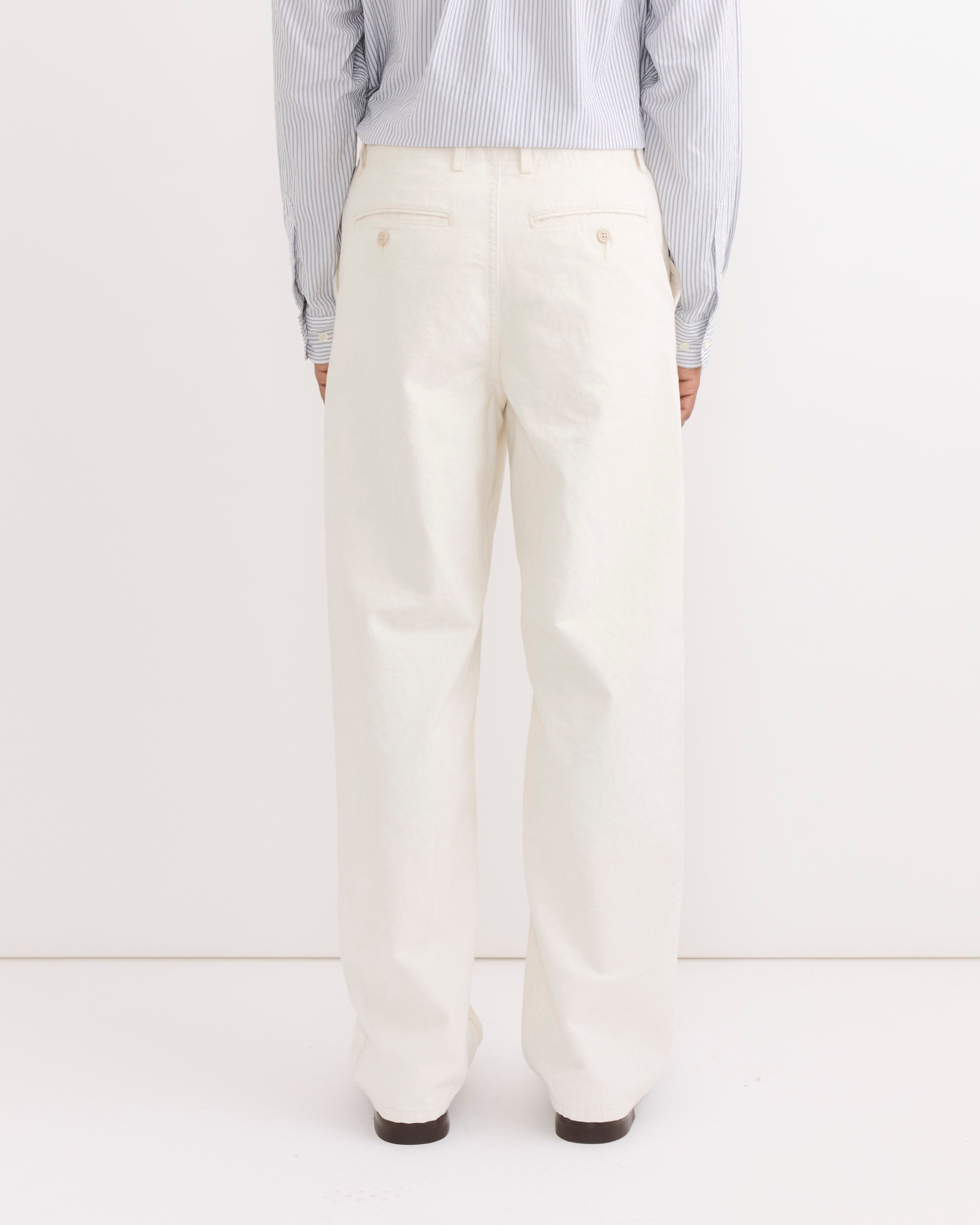 A person stands facing away, wearing SMOCK's Officer Trouser in Washed Pigment Dyed Cotton Natural against a plain white background—ideal for highlighting the fit and style of these chic trousers.