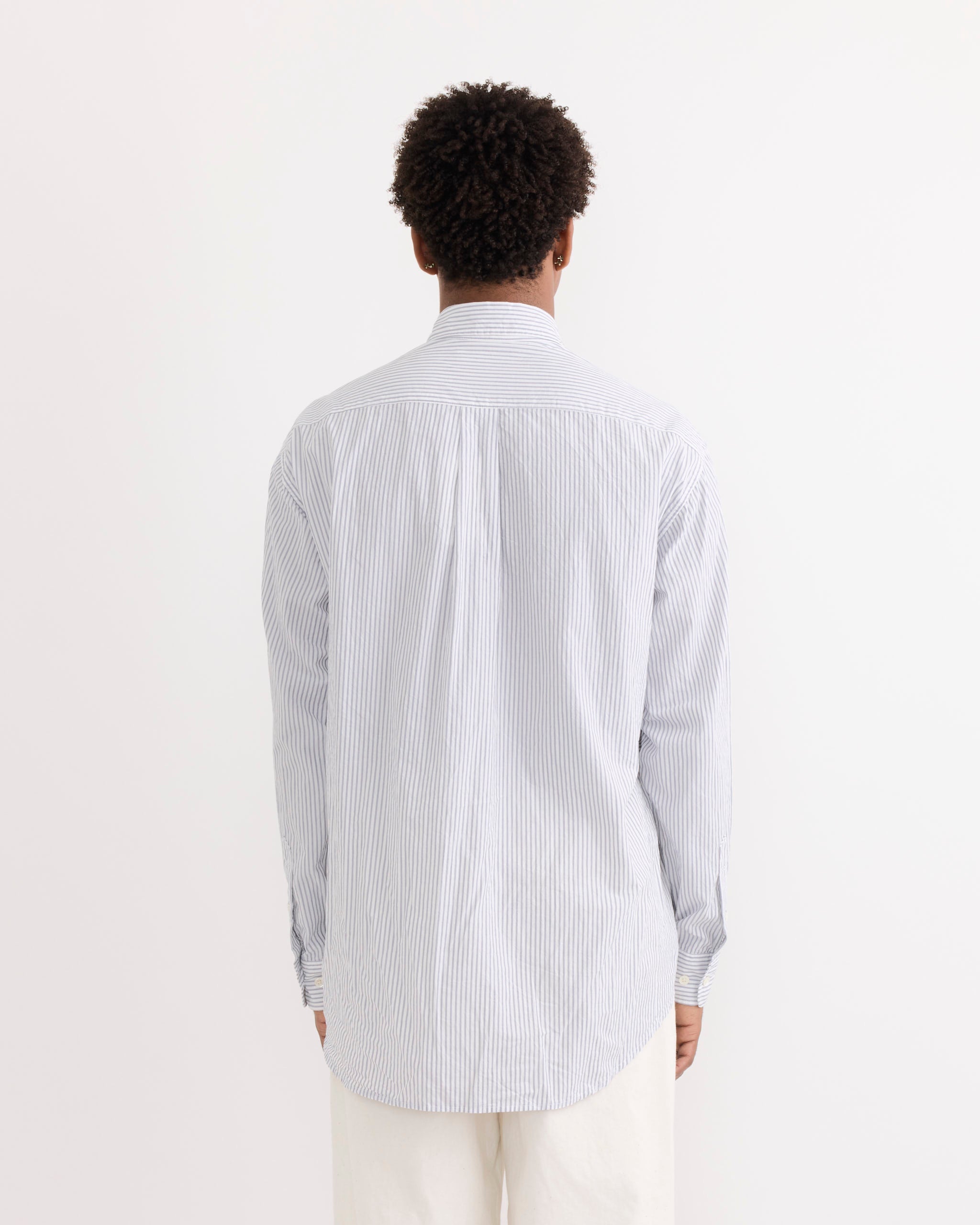 A person with short curly hair faces away, wearing the SMOCK Gio Shirt in Crushed Cotton in Navy Stripe Ni and white pants against a plain white background.