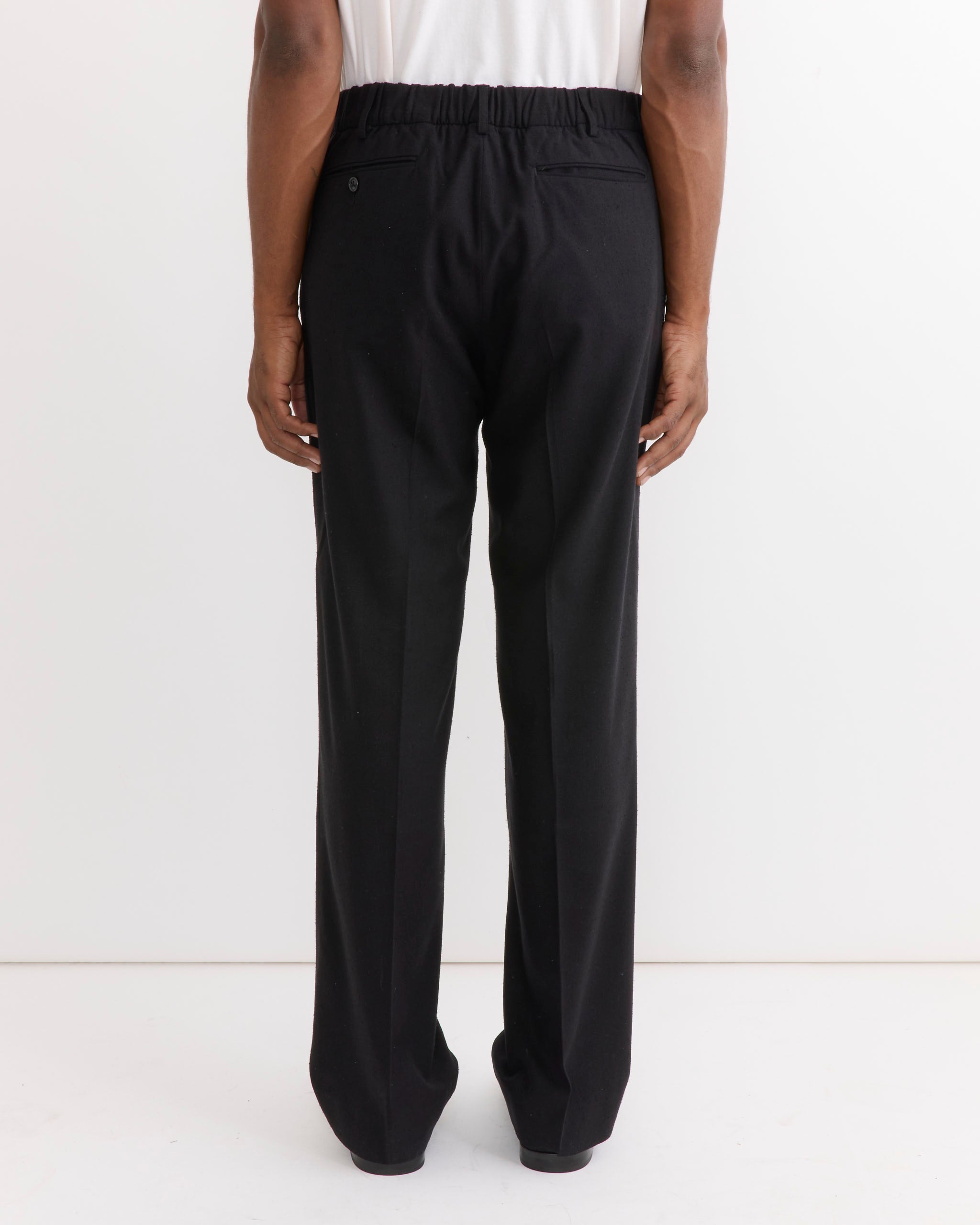 A person stands against a white background, shown from behind, wearing the SMOCK West Coast Pant in Raw Silk Black. The loose-fitting pants feature an elastic waistband and a buttoned pocket on the right side, paired with a white shirt.