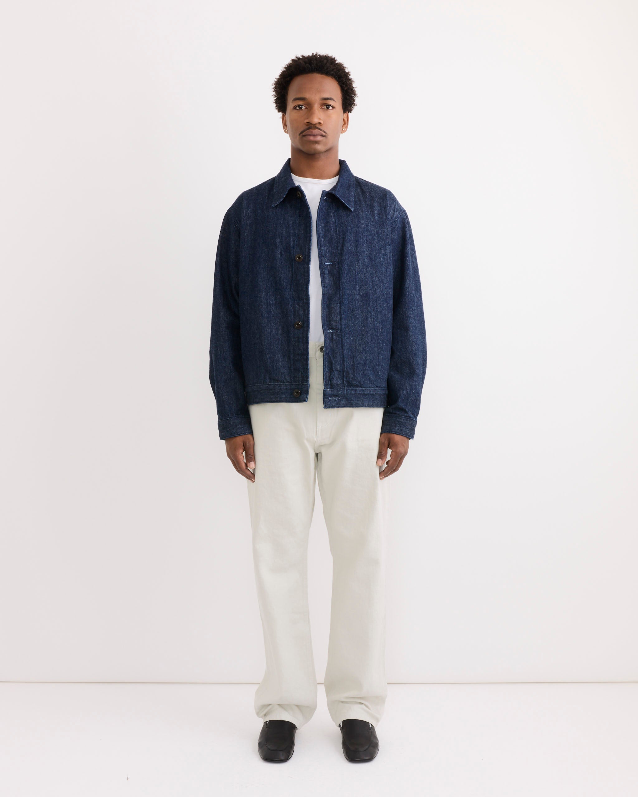 A man stands against a plain white background, wearing the SMOCK Lang Jacket in Selvedge Denim Dark Blue over a light shirt with off-white pants and black shoes. His arms are at his sides, showing fit and sizing. He has a neutral expression.