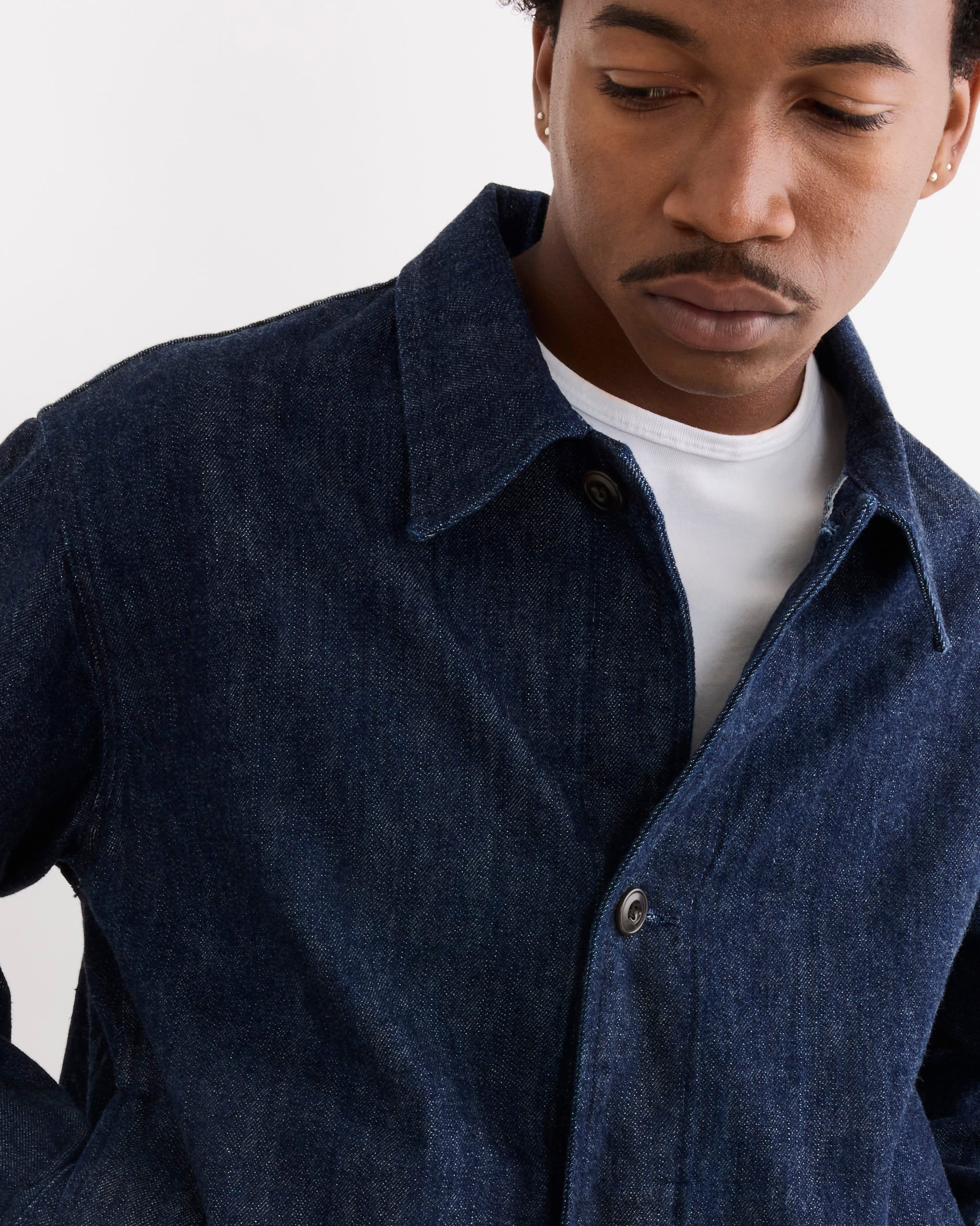 Wearing the SMOCK Lang Jacket in Selvedge Denim Dark Blue over a white shirt, a person looks down against a plain light background, satisfied with the perfect fit and sizing discovered via online chat.