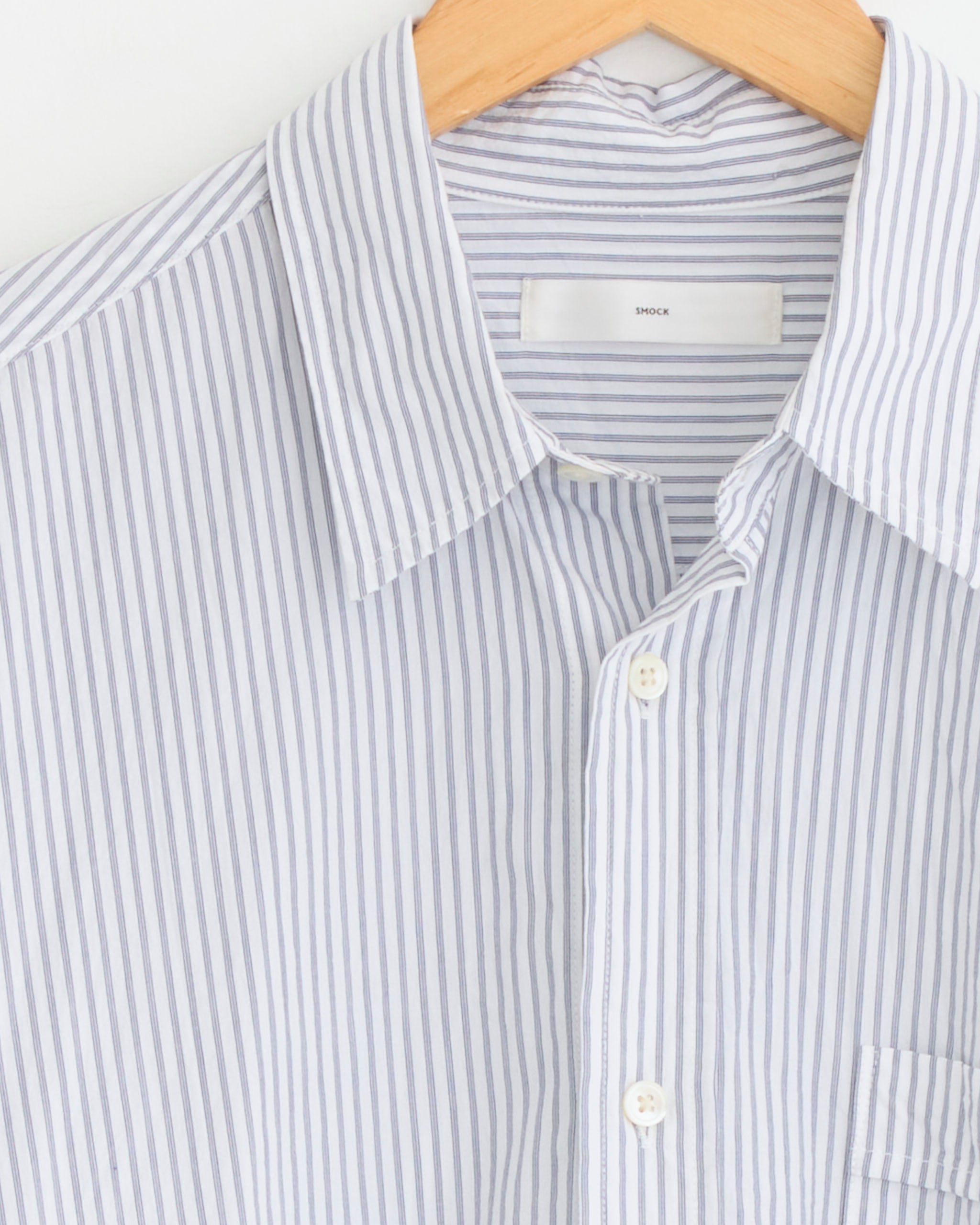 A close-up of the SMOCK Gio Shirt in Crushed Cotton in Navy Stripe Ni, featuring thin blue vertical stripes, a collar, white buttons, and a chest pocket, hanging on a wooden hanger.