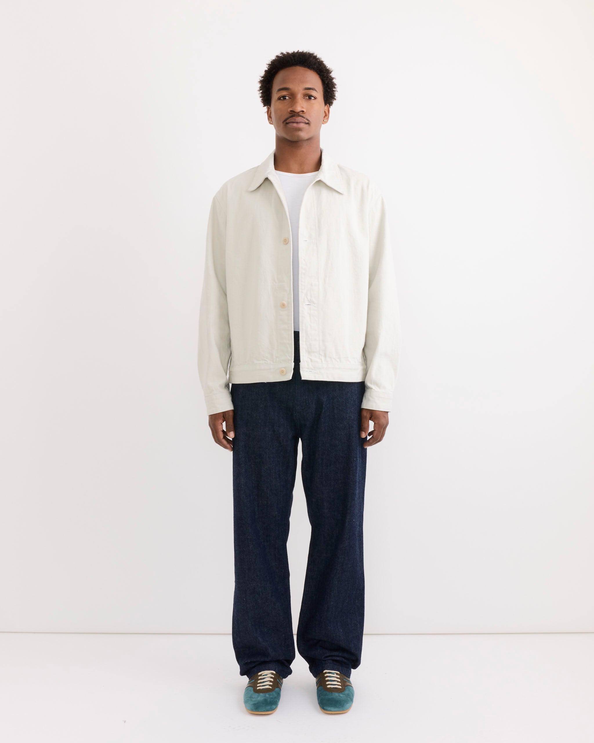A man faces forward against a white background, wearing the SMOCK Lang Jacket in Selvedge Denim Ivory over a white shirt, paired with relaxed dark wide-leg jeans and teal sneakers. For sizing help, contact our online chat.
