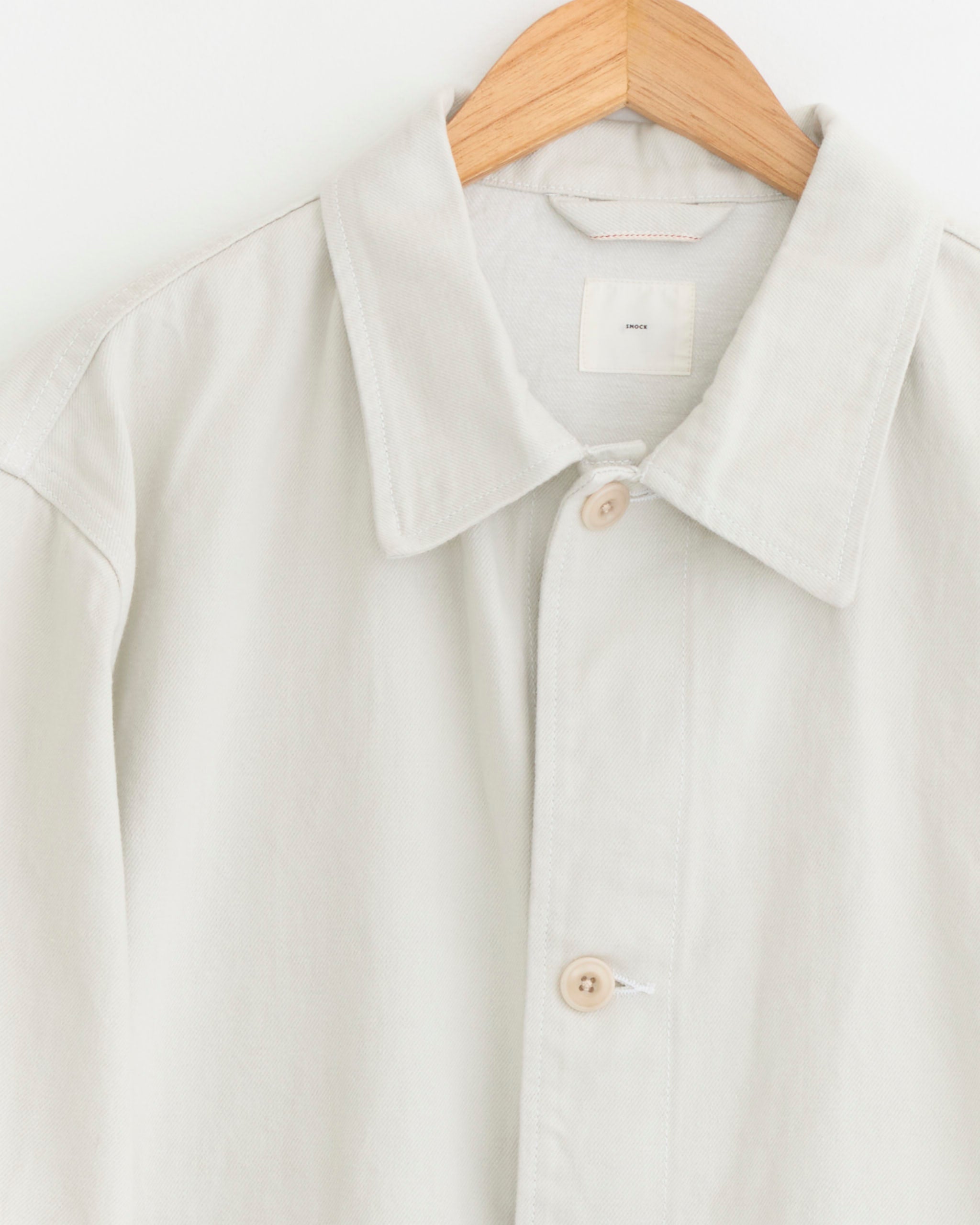 The SMOCK Lang Jacket in Selvedge Denim Ivory, featuring a light beige button-up design with a collar, hangs on a wooden hanger against a white background. For sizing or fit questions, our online chat is here to help.