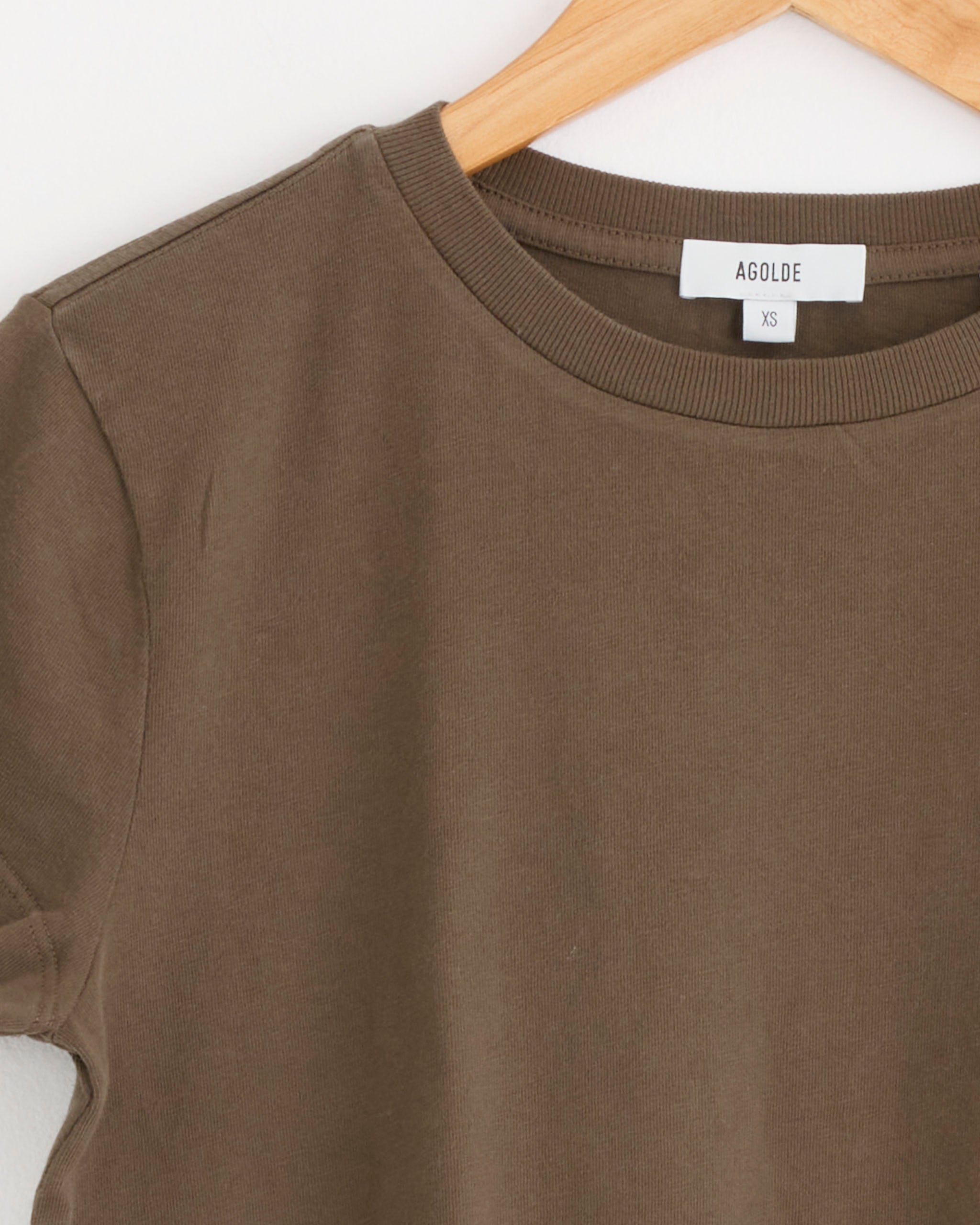 A close-up of the Adine Shrunken Tee in Uniform by AGOLDE, a brown short-sleeve t-shirt in size XS, displayed on a wooden hanger with a visible white AGOLDE tag.