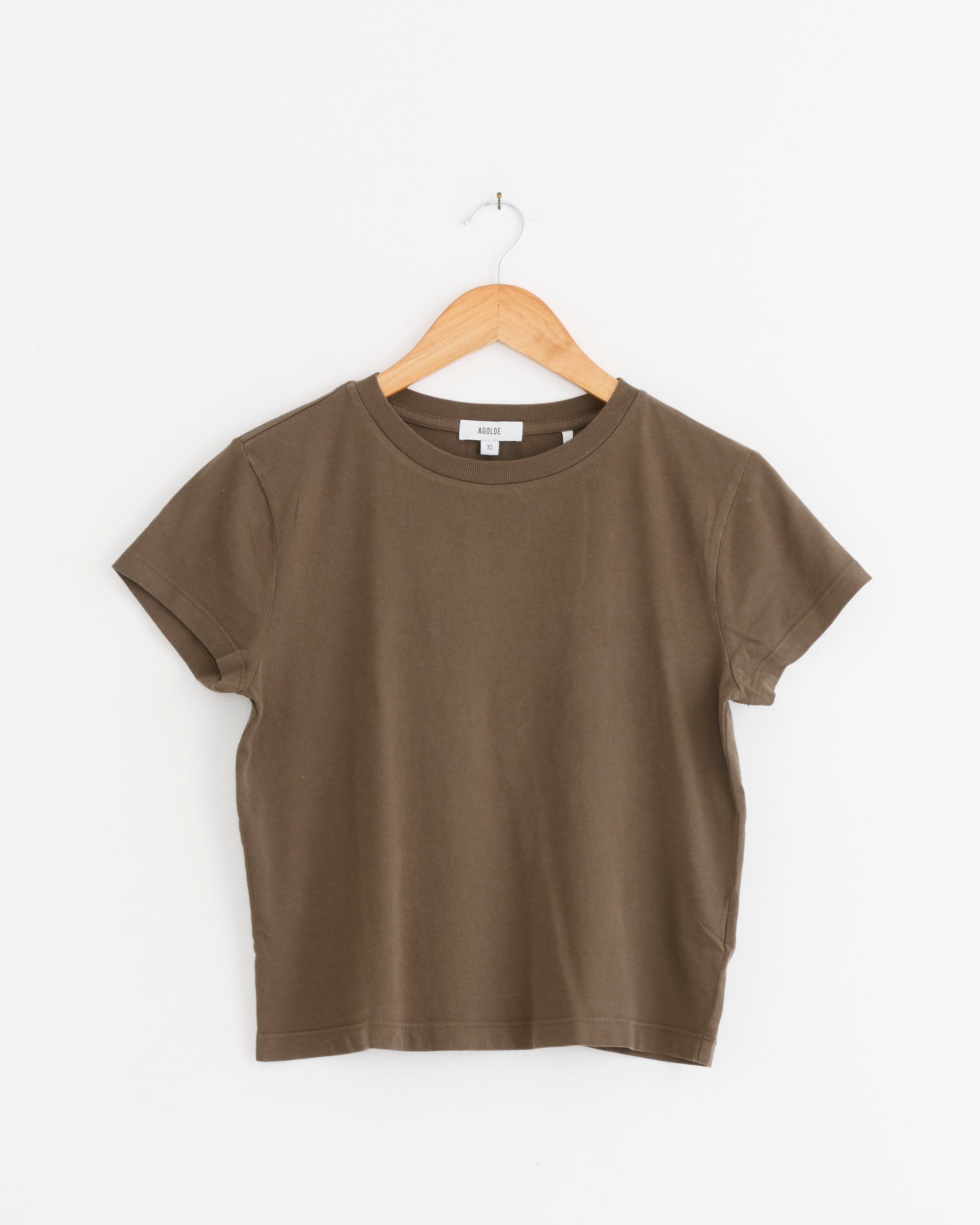 The AGOLDE Adine Shrunken Tee in Uniform, featuring short sleeves and a plain brown design, hangs on a wooden hanger against a white wall.