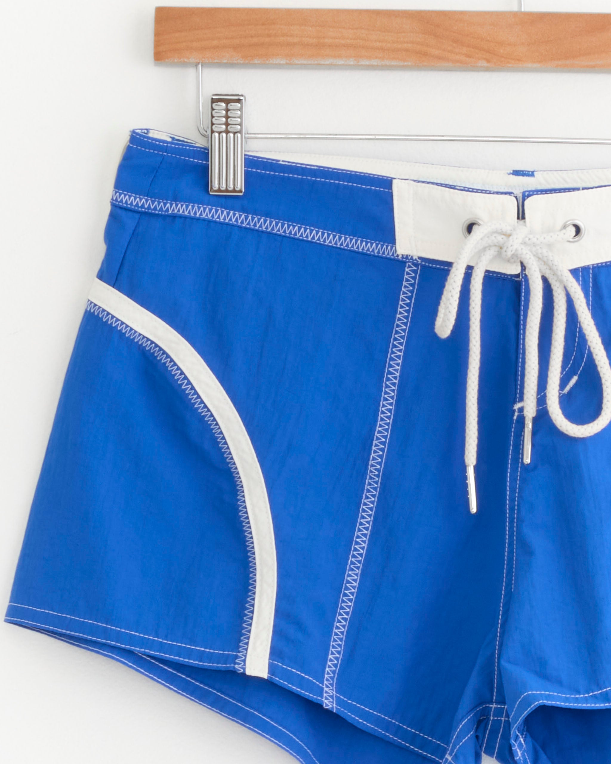 The Short Shorts in Cobalt/White by Made Some Sportswear, featuring crisp white trim and a matching white drawstring, are styled after classic 1970s surf shorts and are displayed on a wooden hanger against a white background.