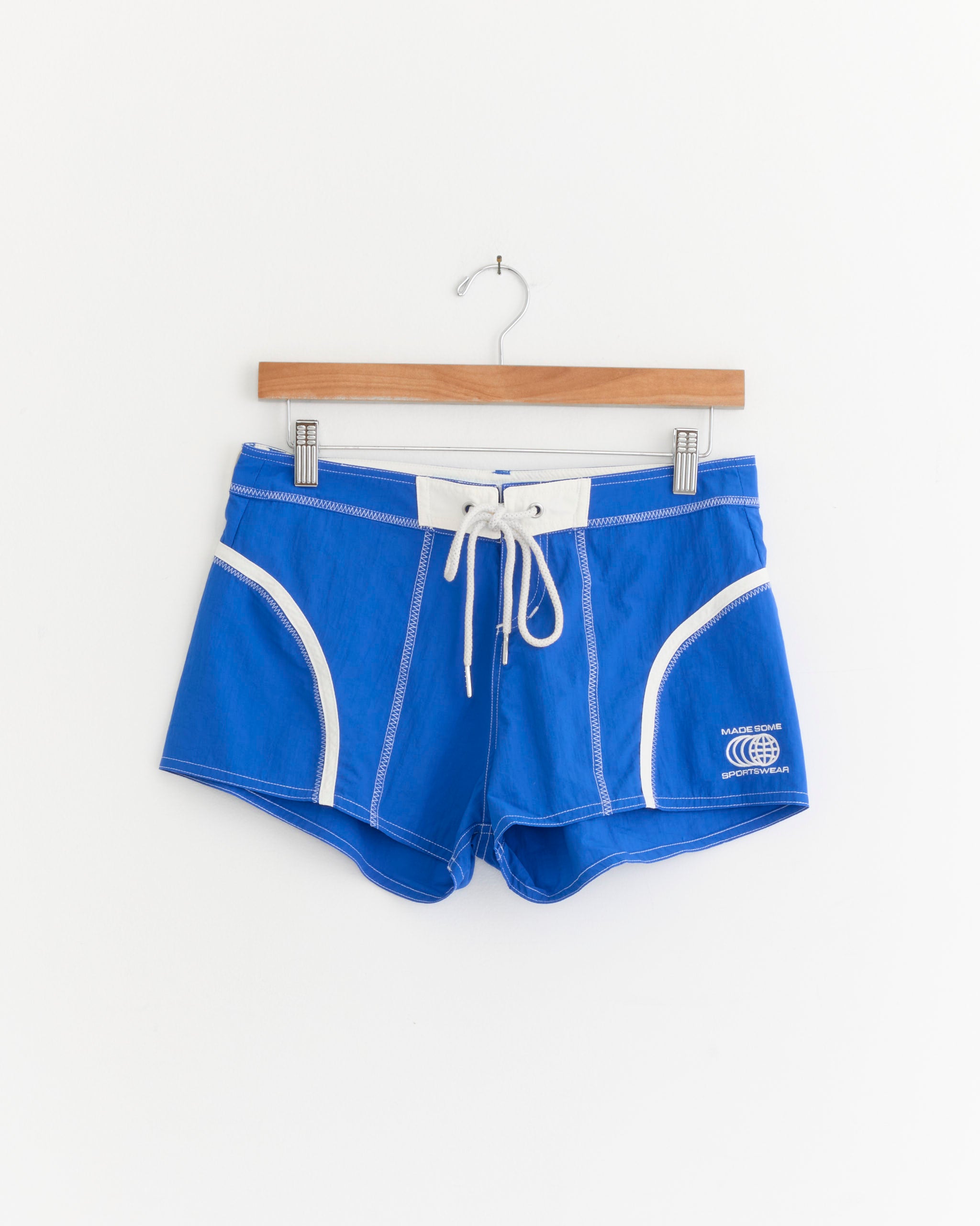 Short Shorts in Cobalt/White by Made Some Sportswear, crafted from lightweight waterproof nylon, feature crisp white trim and a matching drawstring. A small white logo sits near the bottom right hem, evoking classic 1970s surf style. The shorts are displayed on a wooden hanger against a plain white background.