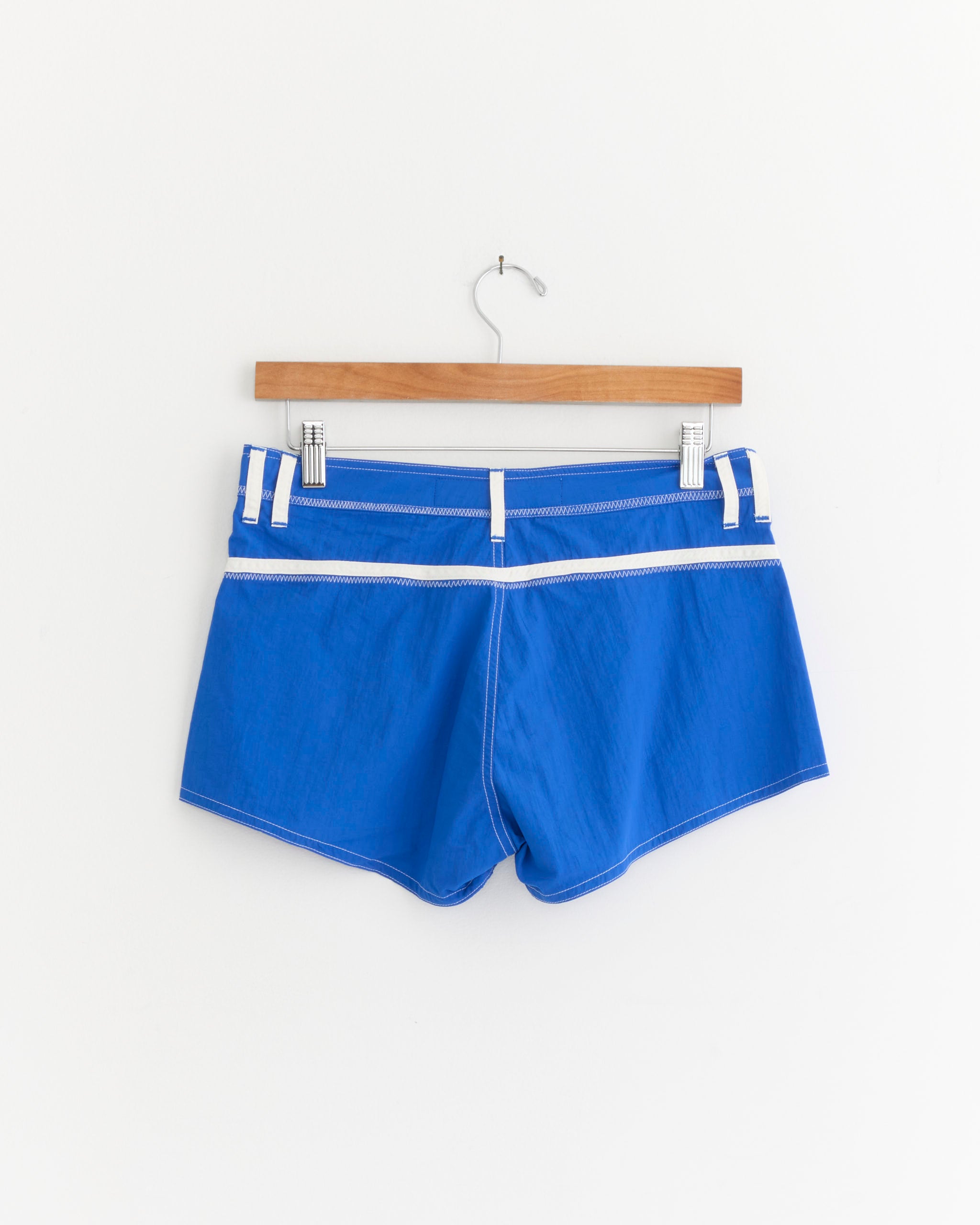 The Short Shorts in Cobalt/White by Made Some Sportswear, inspired by classic 1970s surf shorts, are hanging on a wooden hanger against a plain white background.