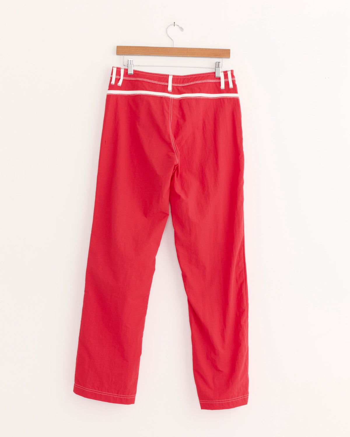 Pant in Red/White by Made Some Sportswear hangs on a wooden hanger against a plain white background. Featuring a vibrant red color with white trim and belt loops, these nylon pants showcase vintage athletic style with their straight-leg design, visible stitching, and lightweight construction—making them ideal for travel.