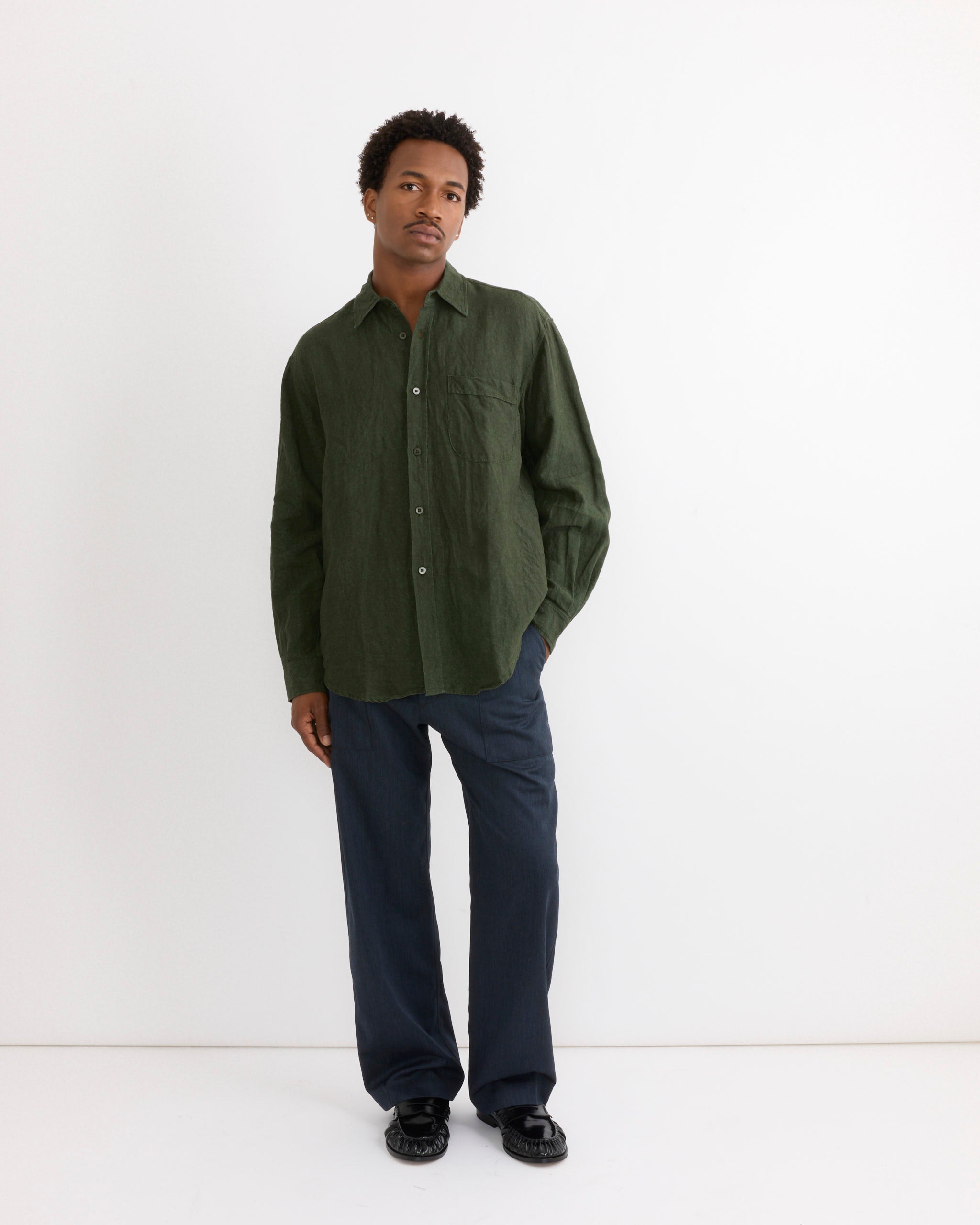 A person stands against a plain white wall, wearing the SMOCK Gio Shirt in Sun Dried Linen Green—crafted from Japanese linen and made in Los Angeles—paired with dark blue trousers and black shoes, with one hand in their pocket.