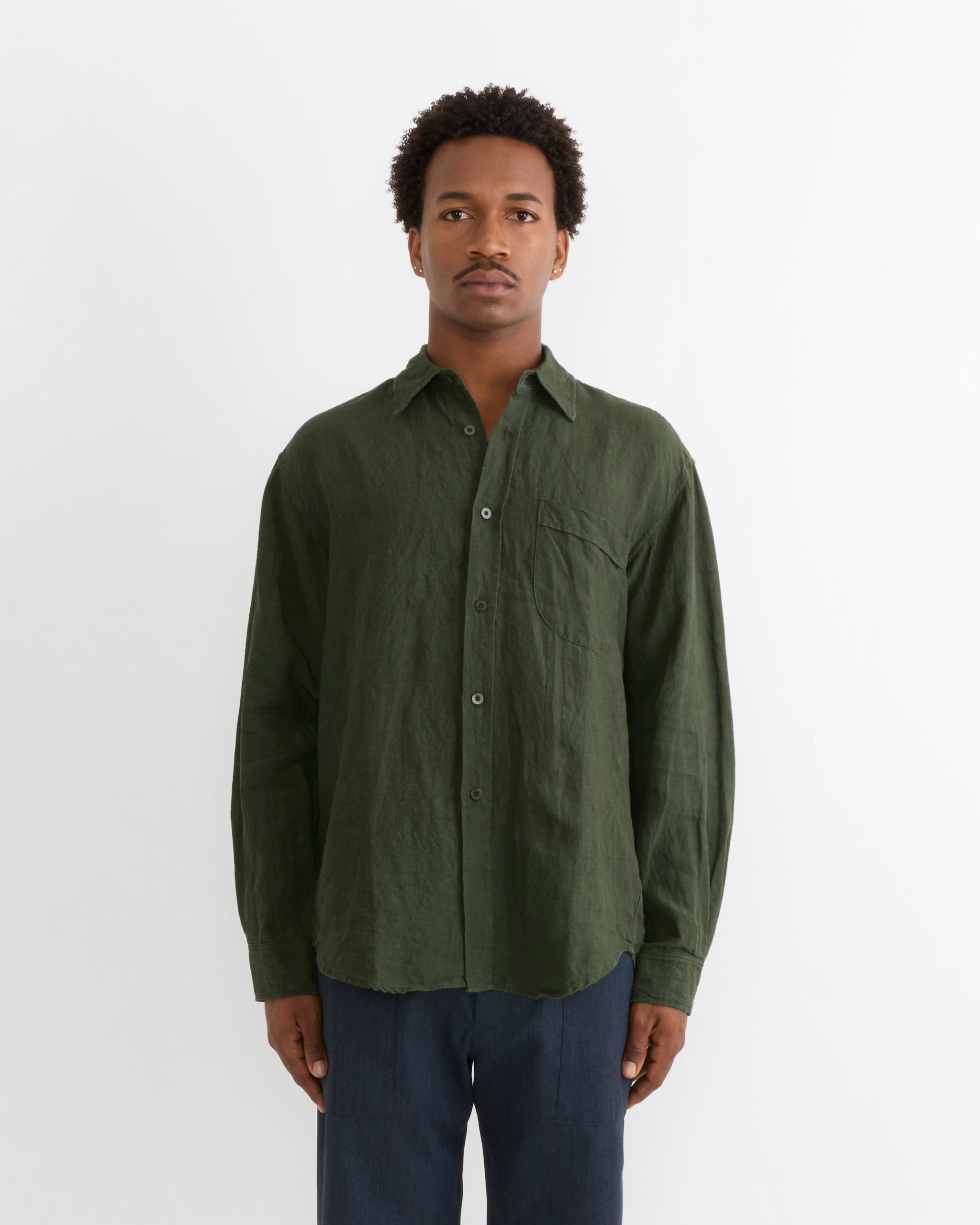 A person stands against a plain white background, wearing the SMOCK Gio Shirt in Sun Dried Linen Green with dark pants, looking straight ahead with a neutral expression.