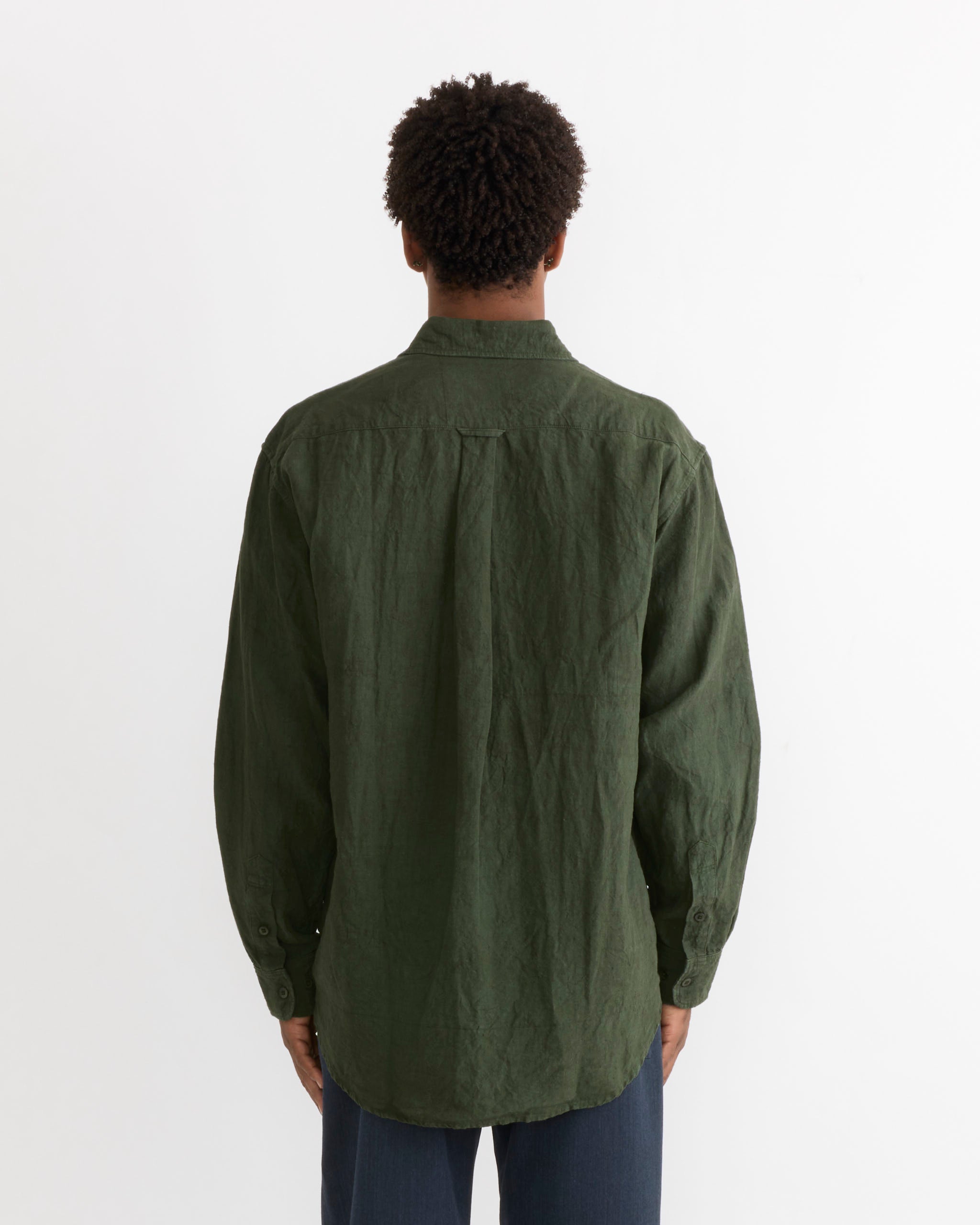 A person stands facing away from the camera, wearing SMOCK’s Gio Shirt in Sun Dried Linen Green—a loose-fitting, long-sleeve Japanese linen shirt—paired with dark pants against a plain white background. This thoughtfully made piece showcases the relaxed elegance of sun dried linen and is crafted in Los Angeles.